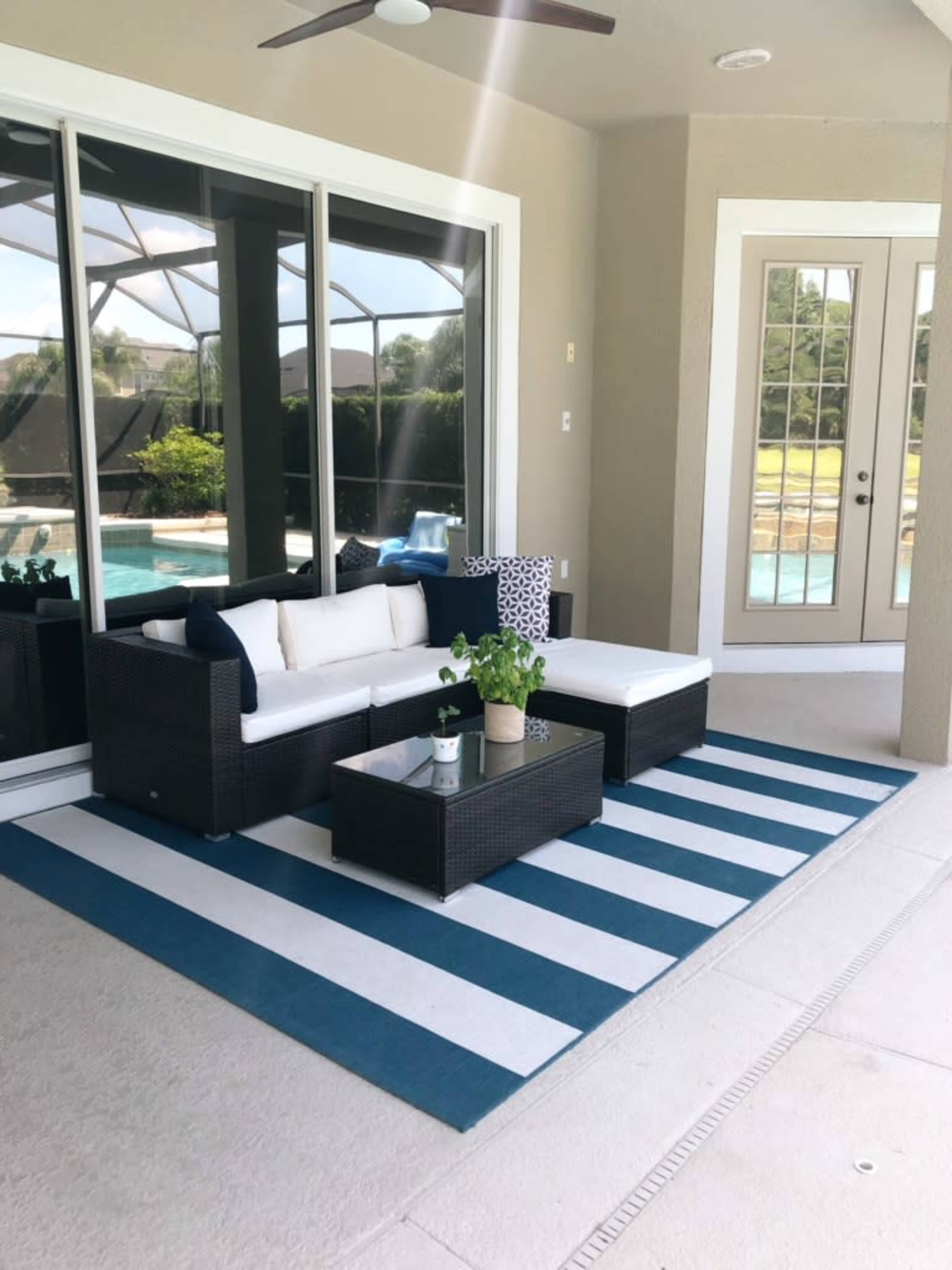 A patio area with a white and blue striped rug, a wicker seating arrangement, and a small table with a plant, all facing a large window overlooking a pool.