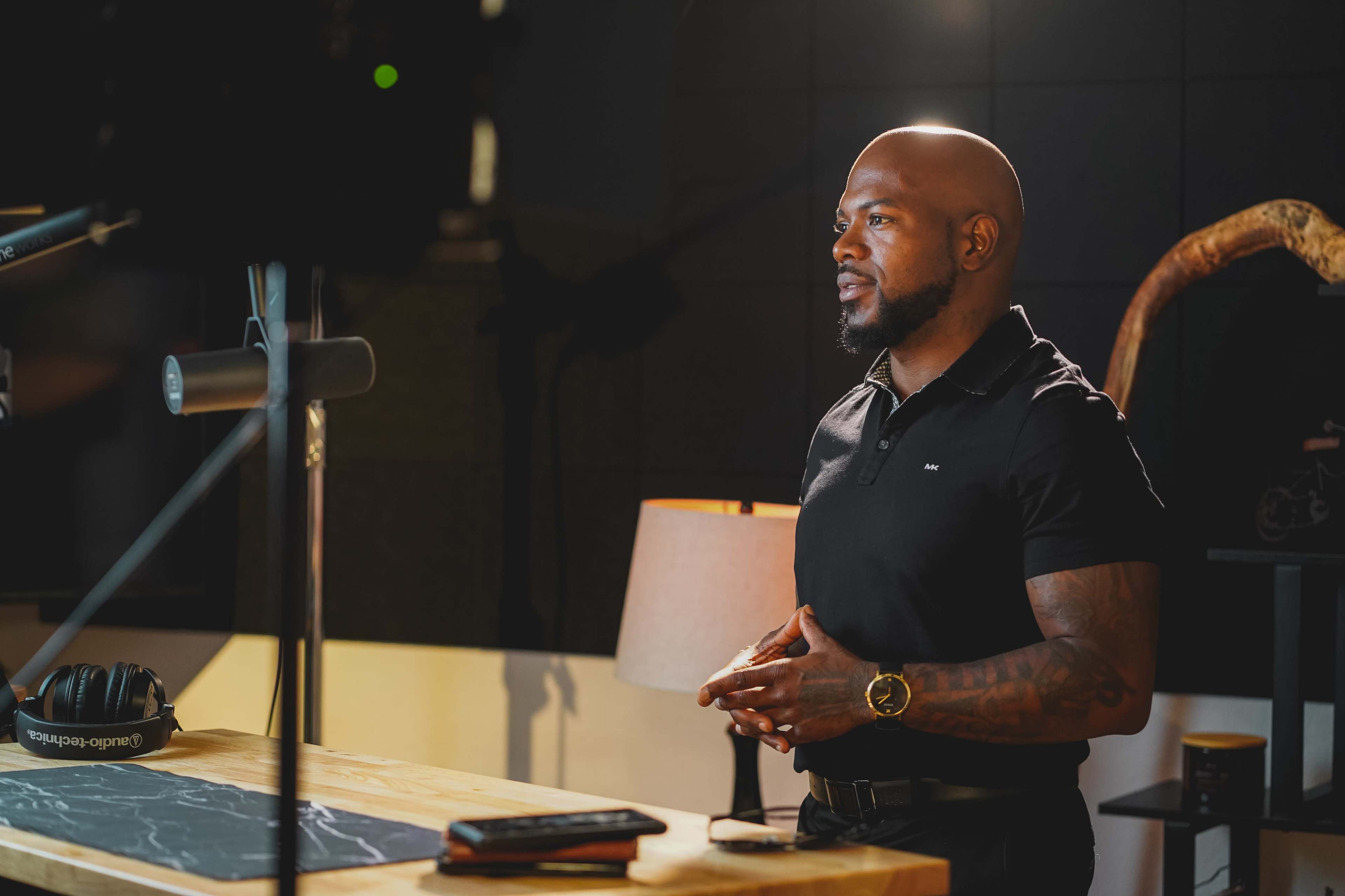 A man stands at a wooden table in a well-lit studio setting, wearing a black polo shirt and a watch, with a microphone and lamp nearby.