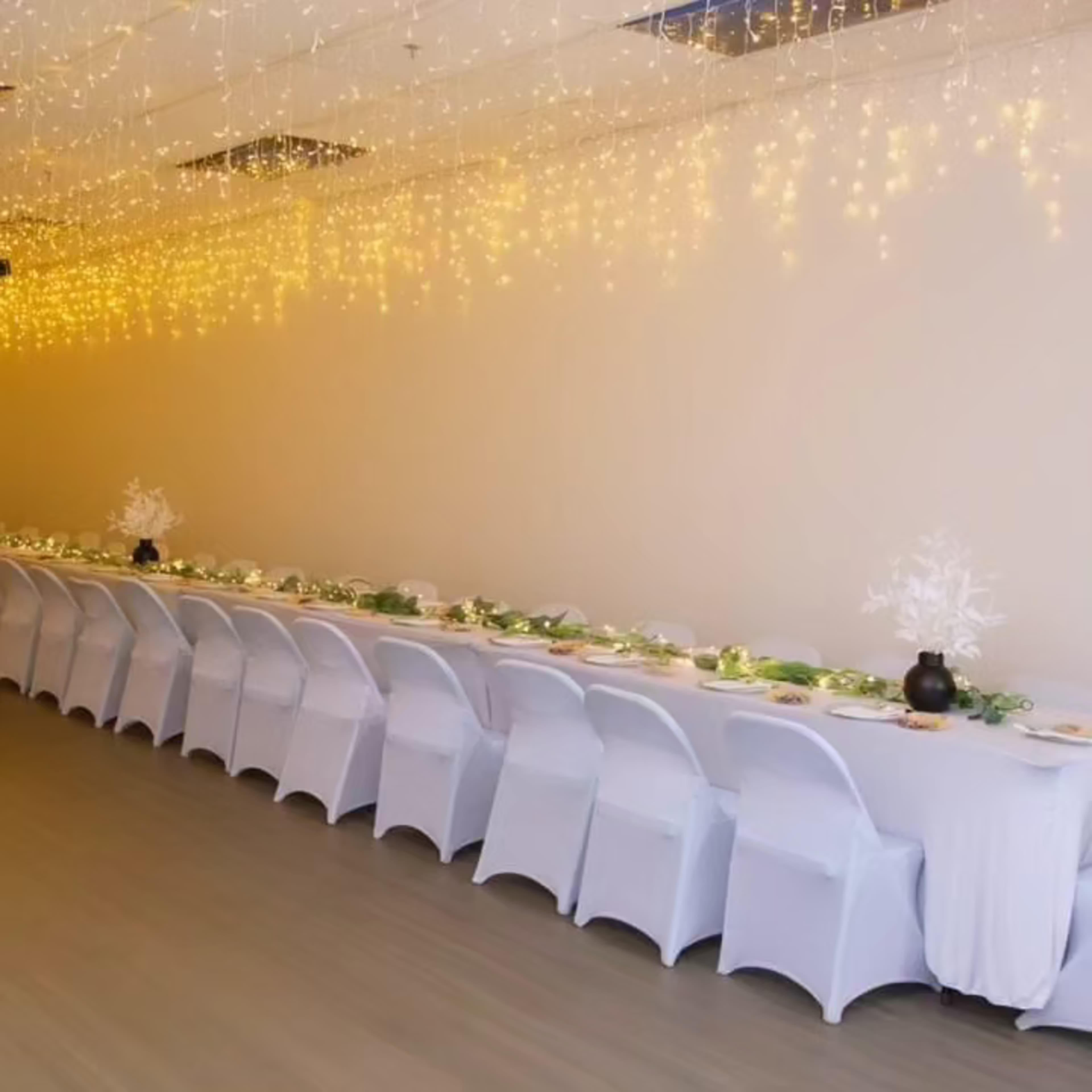 A long white table is set with dishes, surrounded by white chairs, beneath strings of warm fairy lights.