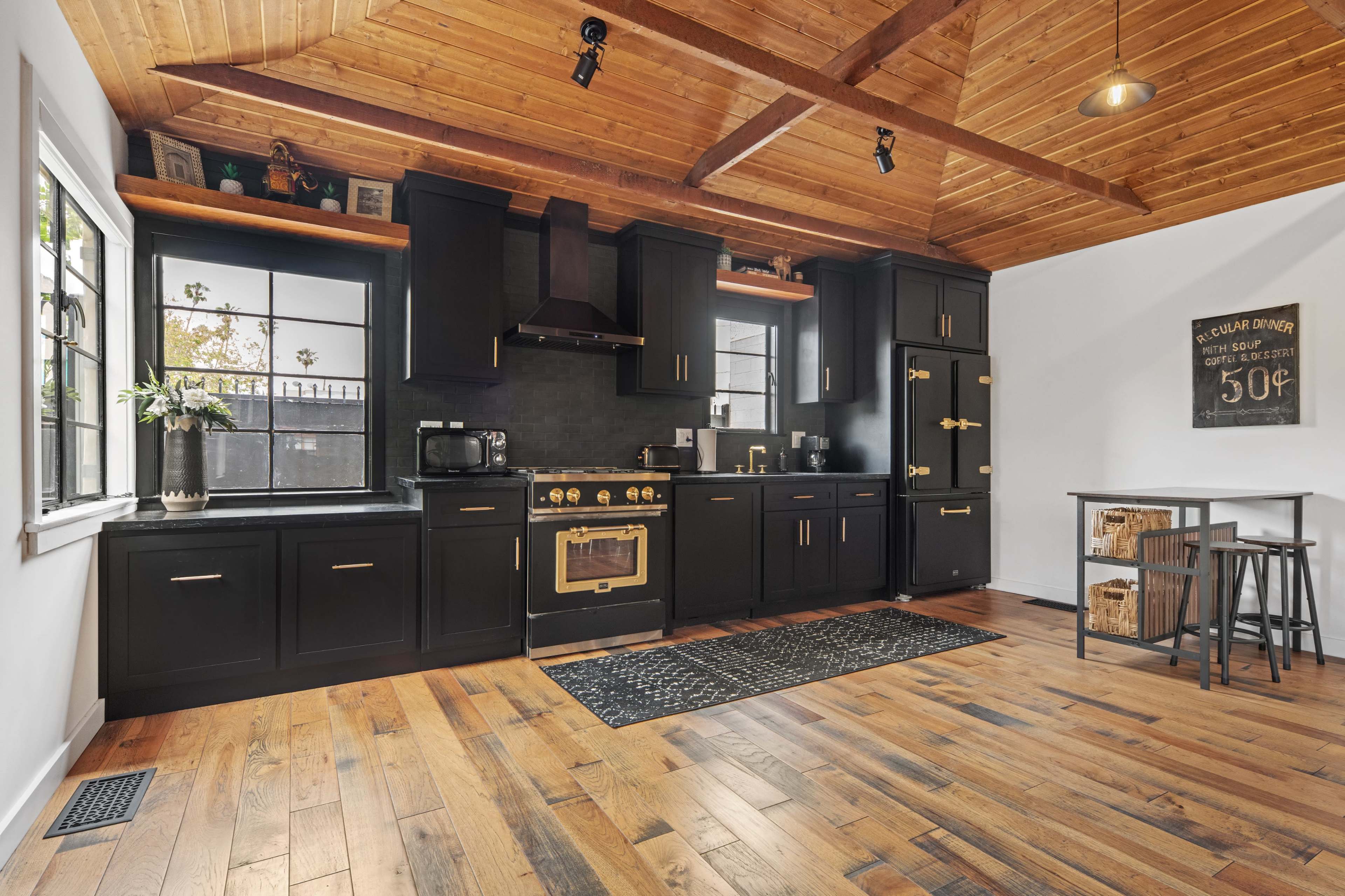 A modern kitchen with black cabinetry, wooden beams ceiling, and a wooden floor, featuring stainless steel appliances and a dining area with stools.