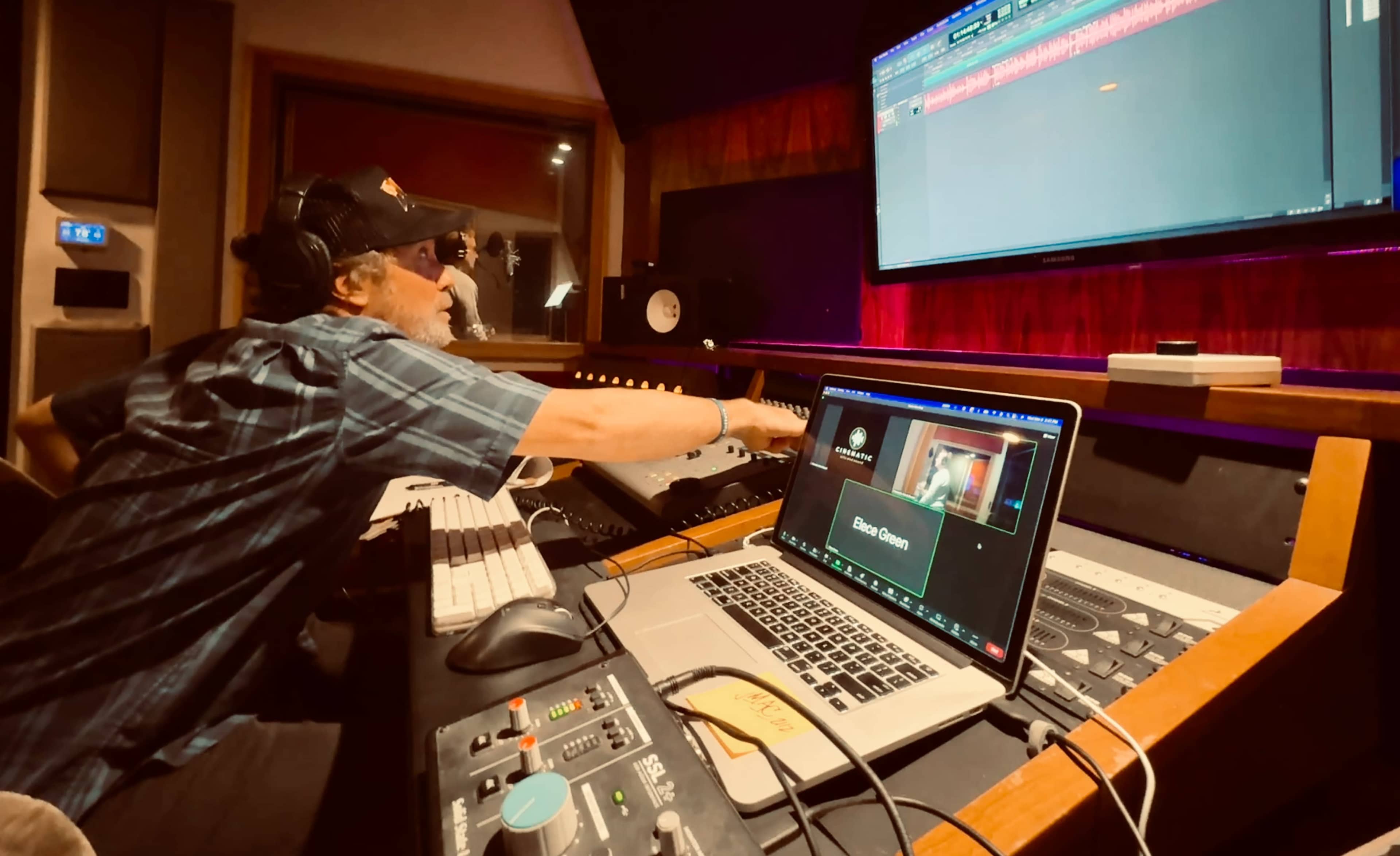 A person is working in a music studio, adjusting controls on a console while looking at a computer screen displaying audio tracks.