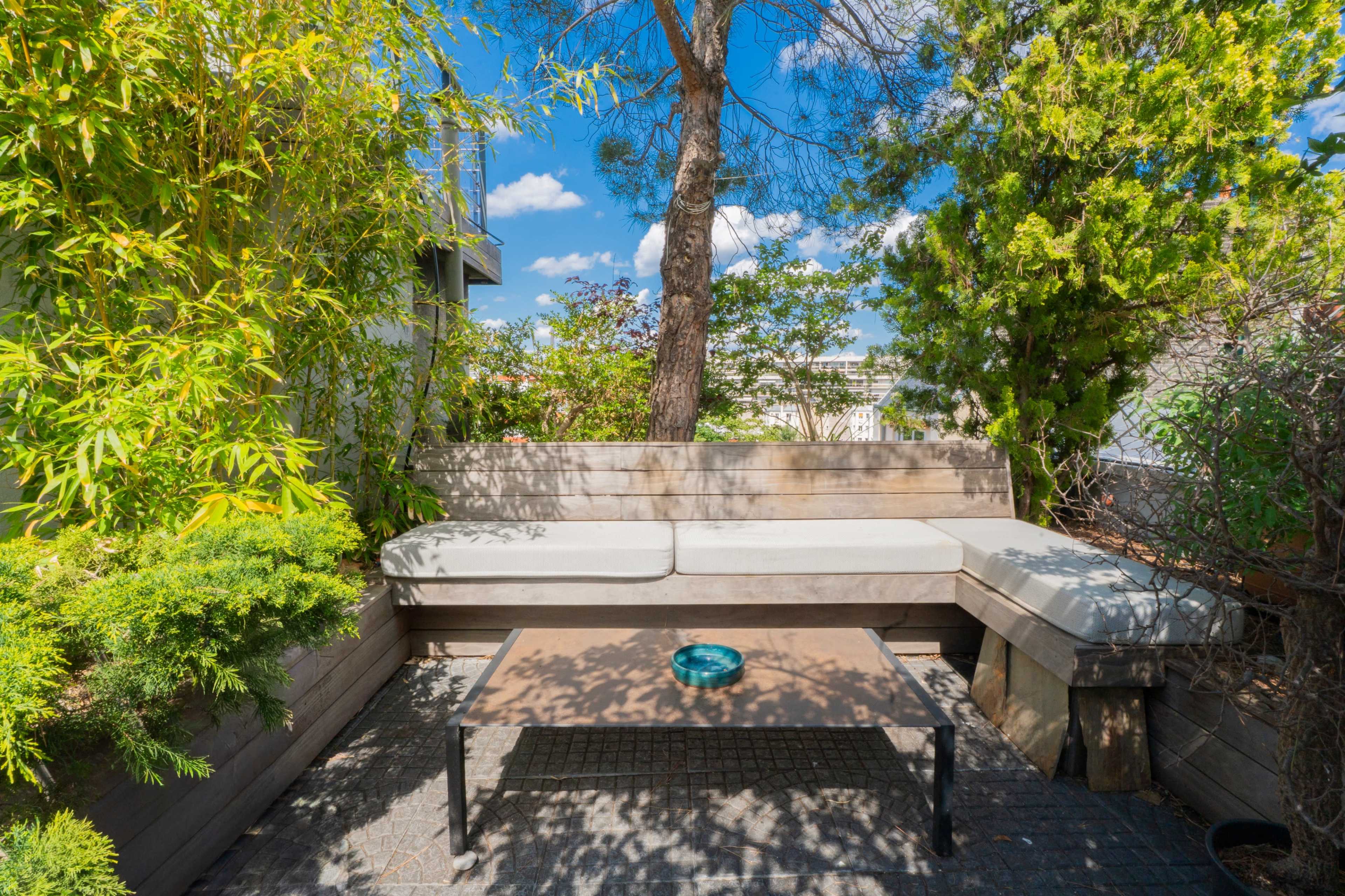 The image shows a small outdoor seating area with a wooden bench, a coffee table, and surrounding greenery under a blue sky.