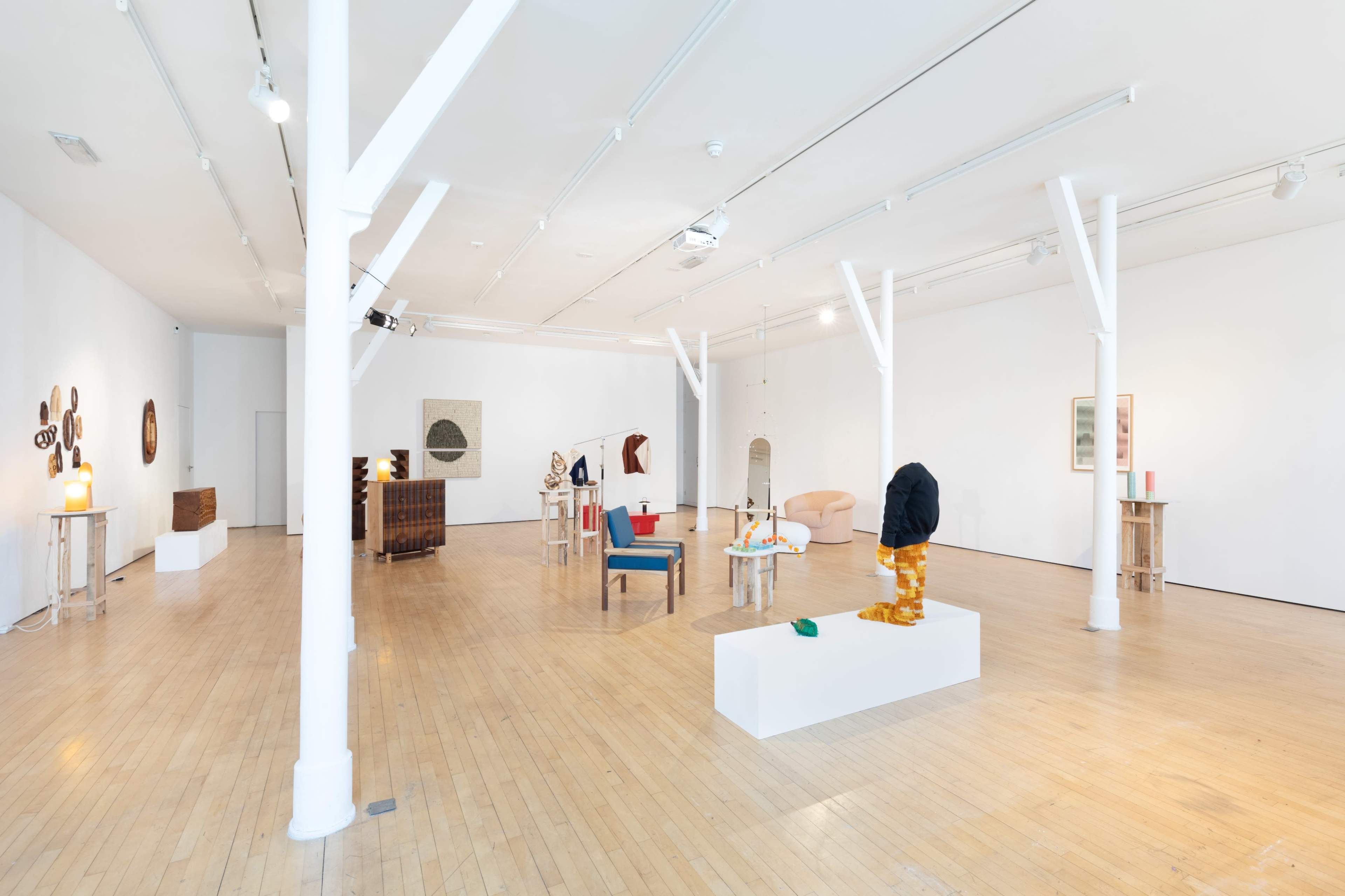A spacious art gallery with wooden floors and white walls, featuring various sculptures and artworks displayed on pedestals and furniture throughout the room.