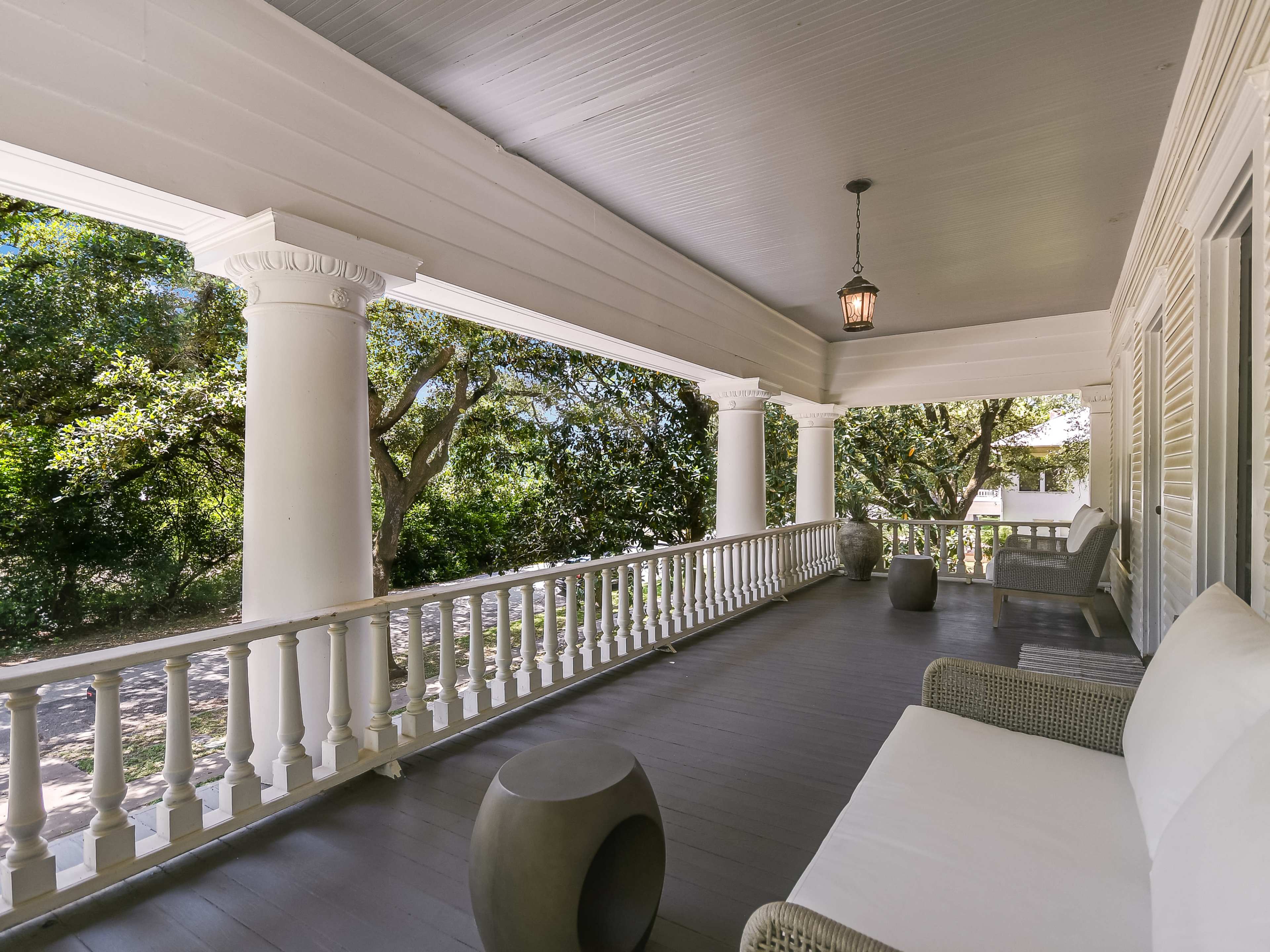 A porch with white columns and seating areas, surrounded by green trees.
