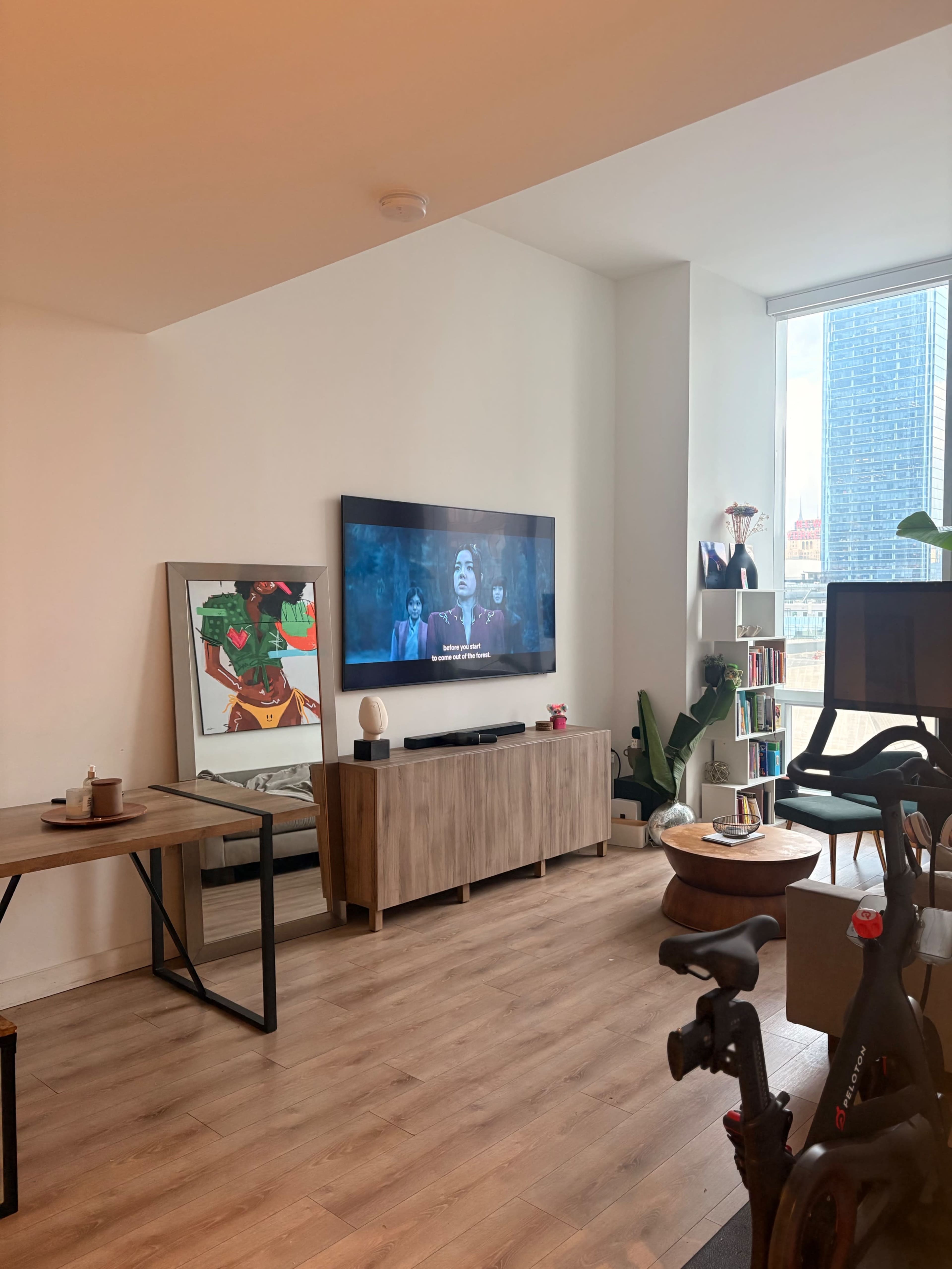 A modern living room features a wall-mounted TV displaying a scene from a movie, a wooden sideboard, a table with coffee cups, and large windows letting in natural light.