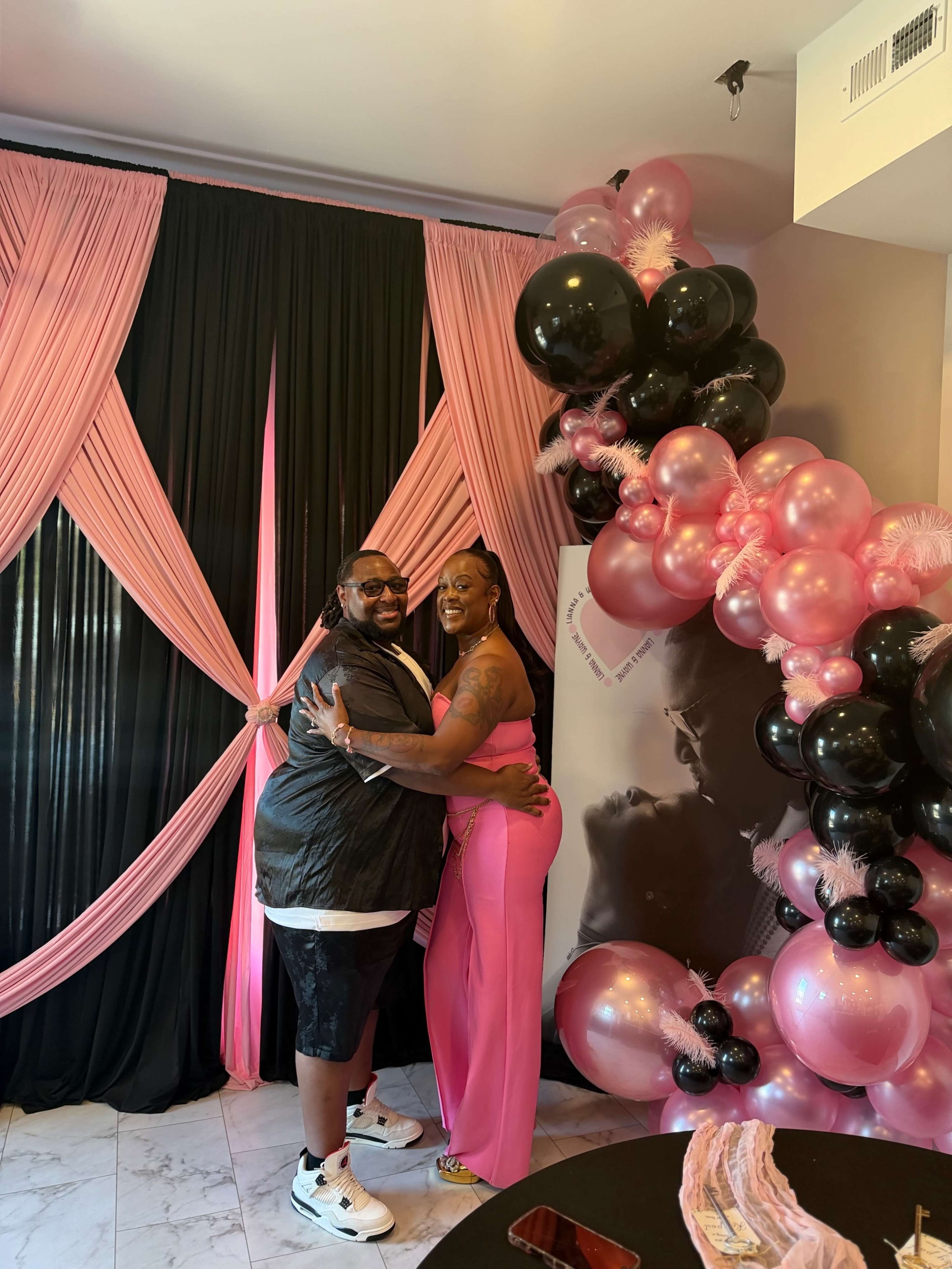 A man and a woman pose together in front of a backdrop of black and pink drapes, surrounded by a balloon arch of various shades of pink and black.