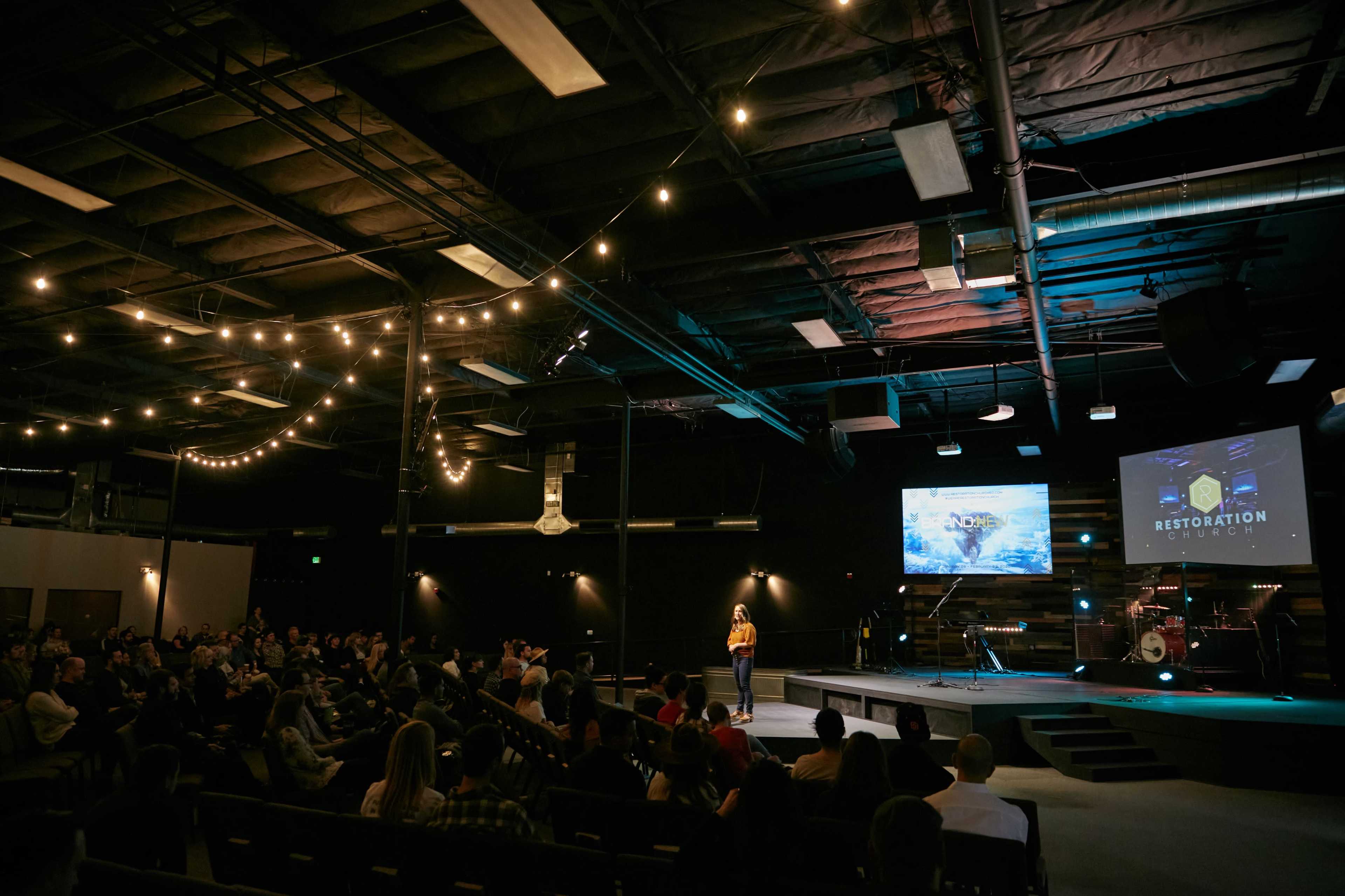 A speaker addresses an audience in a well-lit auditorium featuring a stage with screens displaying graphics.