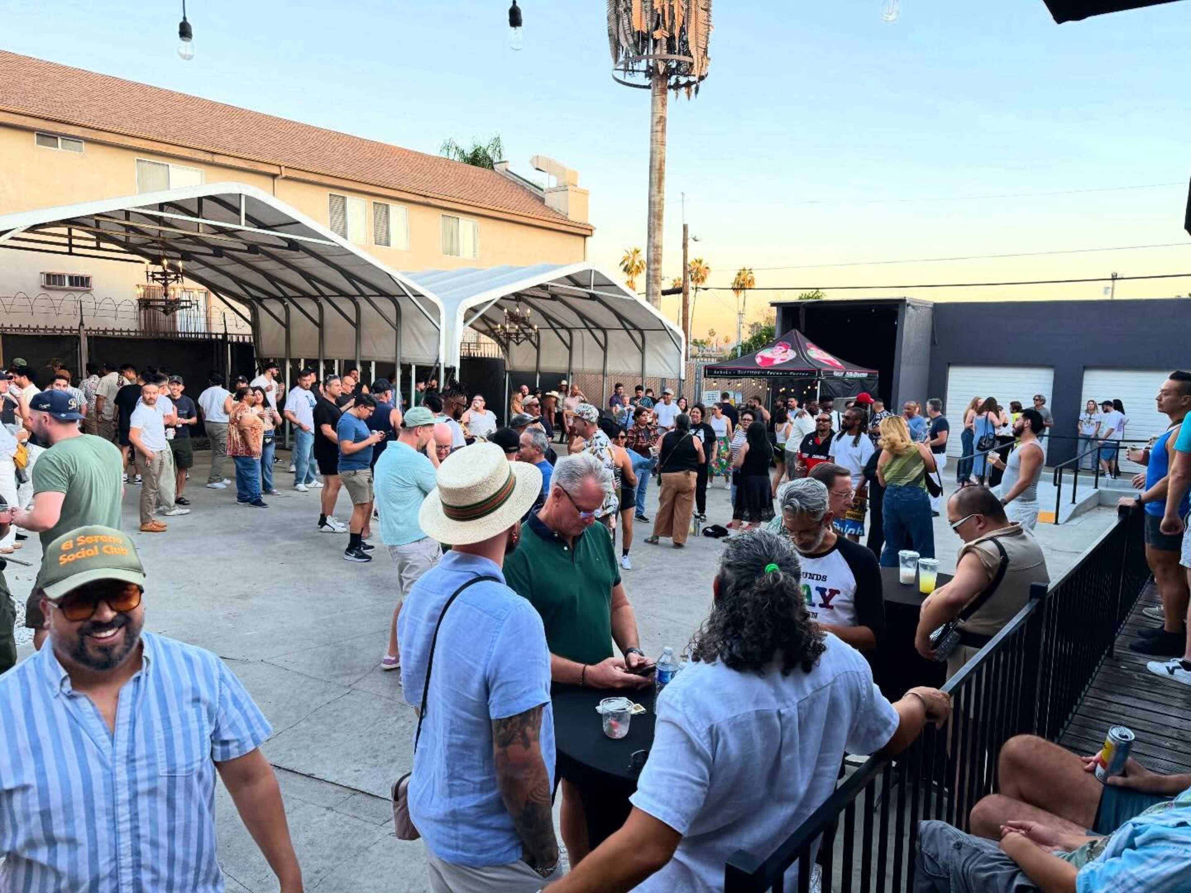 A large crowd of people gathers at an outdoor venue with a tented area and a bar, enjoying drinks and socializing.