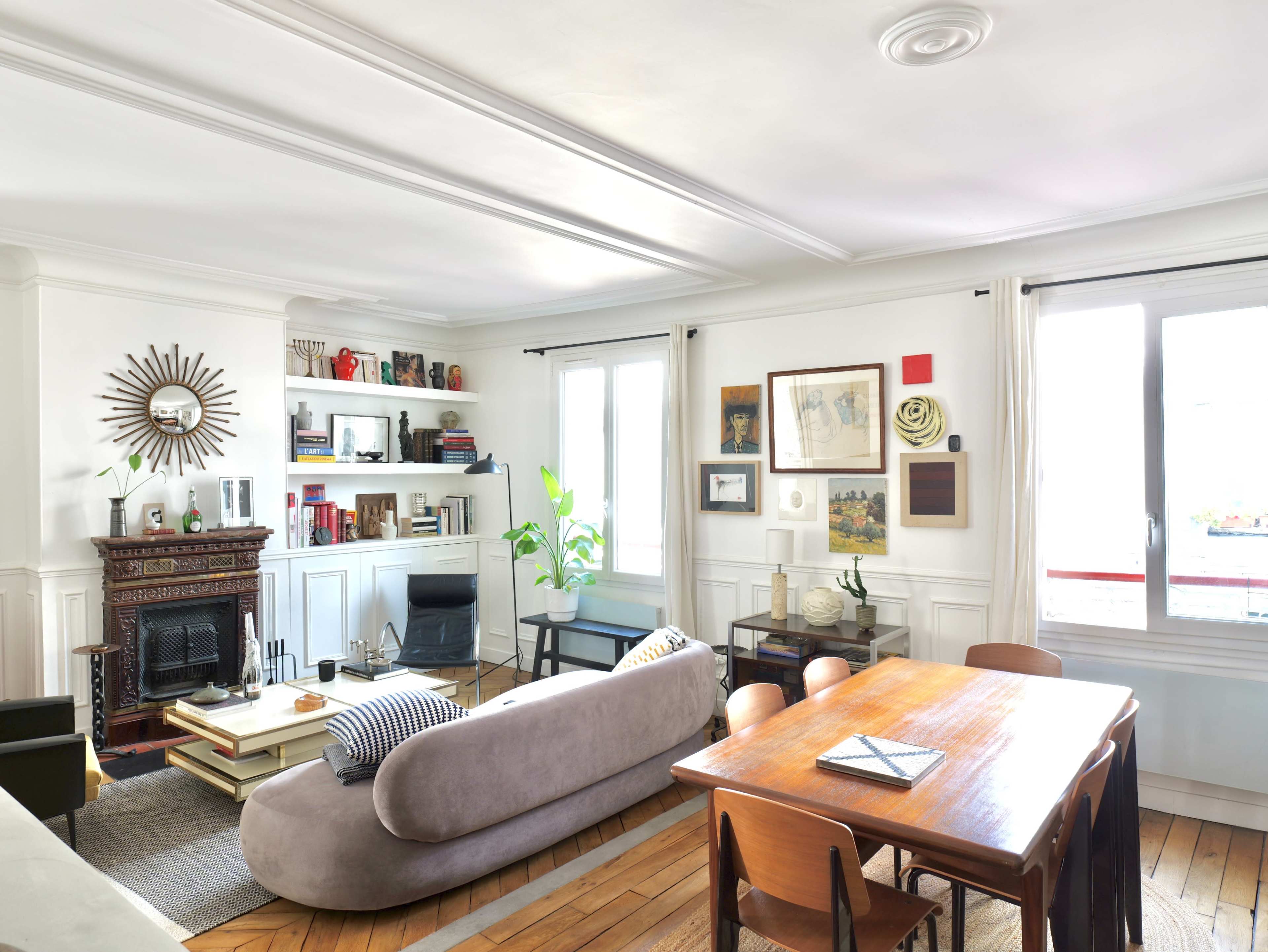 The image shows a bright living room with a dining area, featuring a fireplace, bookshelves, and large windows.