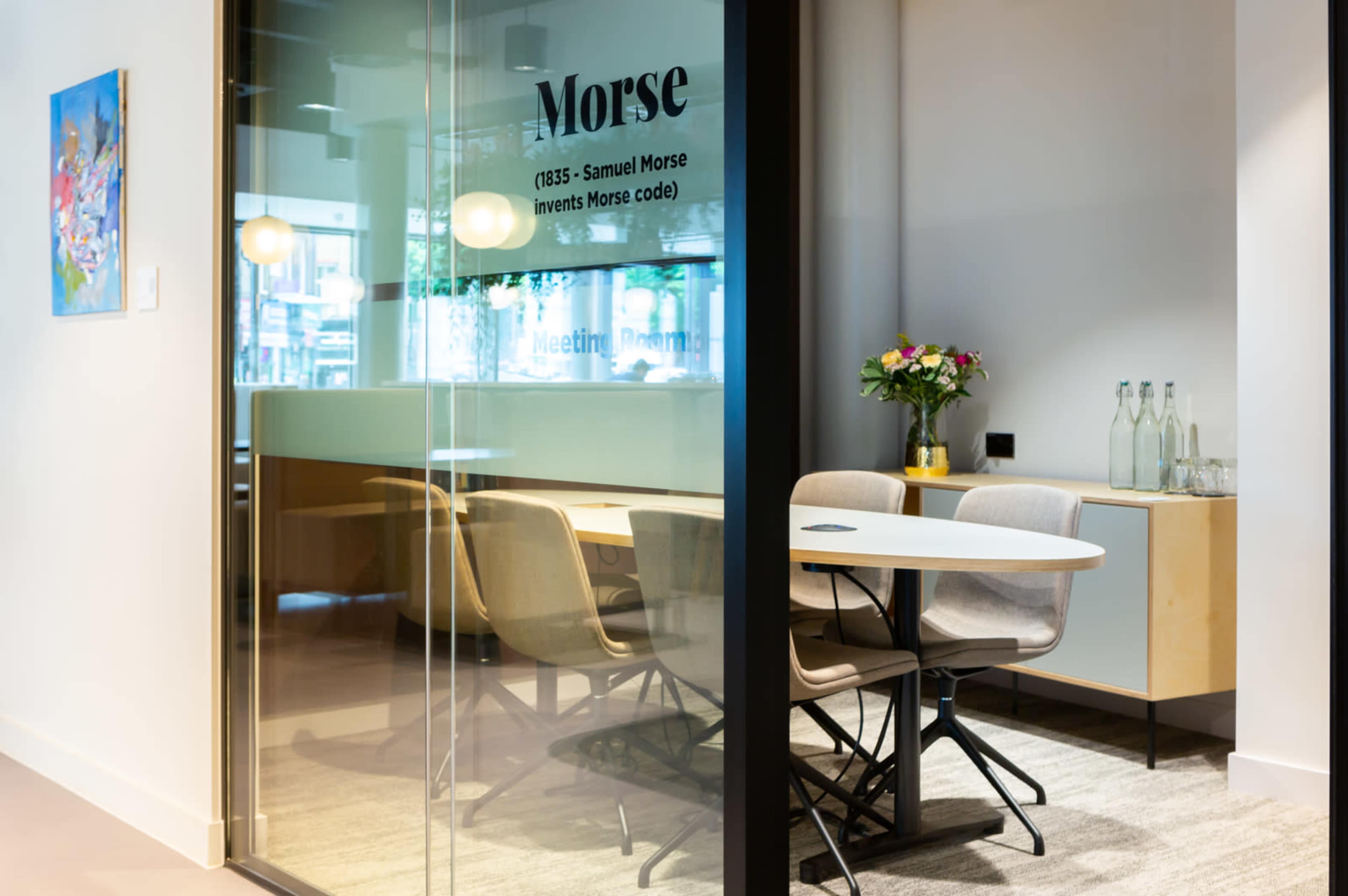 A glass-walled meeting room named "Morse" features a table and chairs, with a decorative bouquet and bottles on a side cabinet.