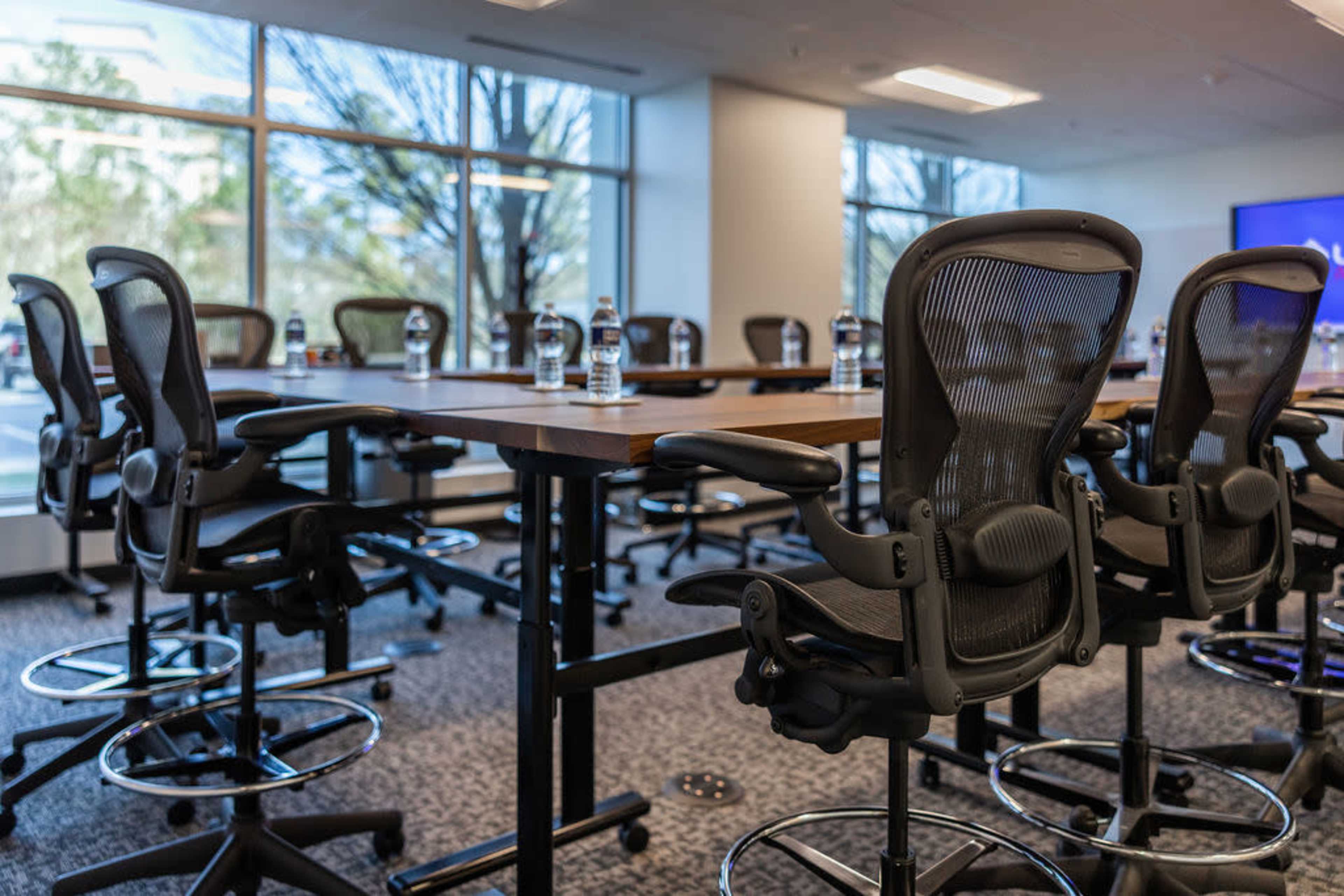 A conference room features several black ergonomic chairs arranged around a long wooden table, with water bottles placed on the table and large windows allowing natural light.
