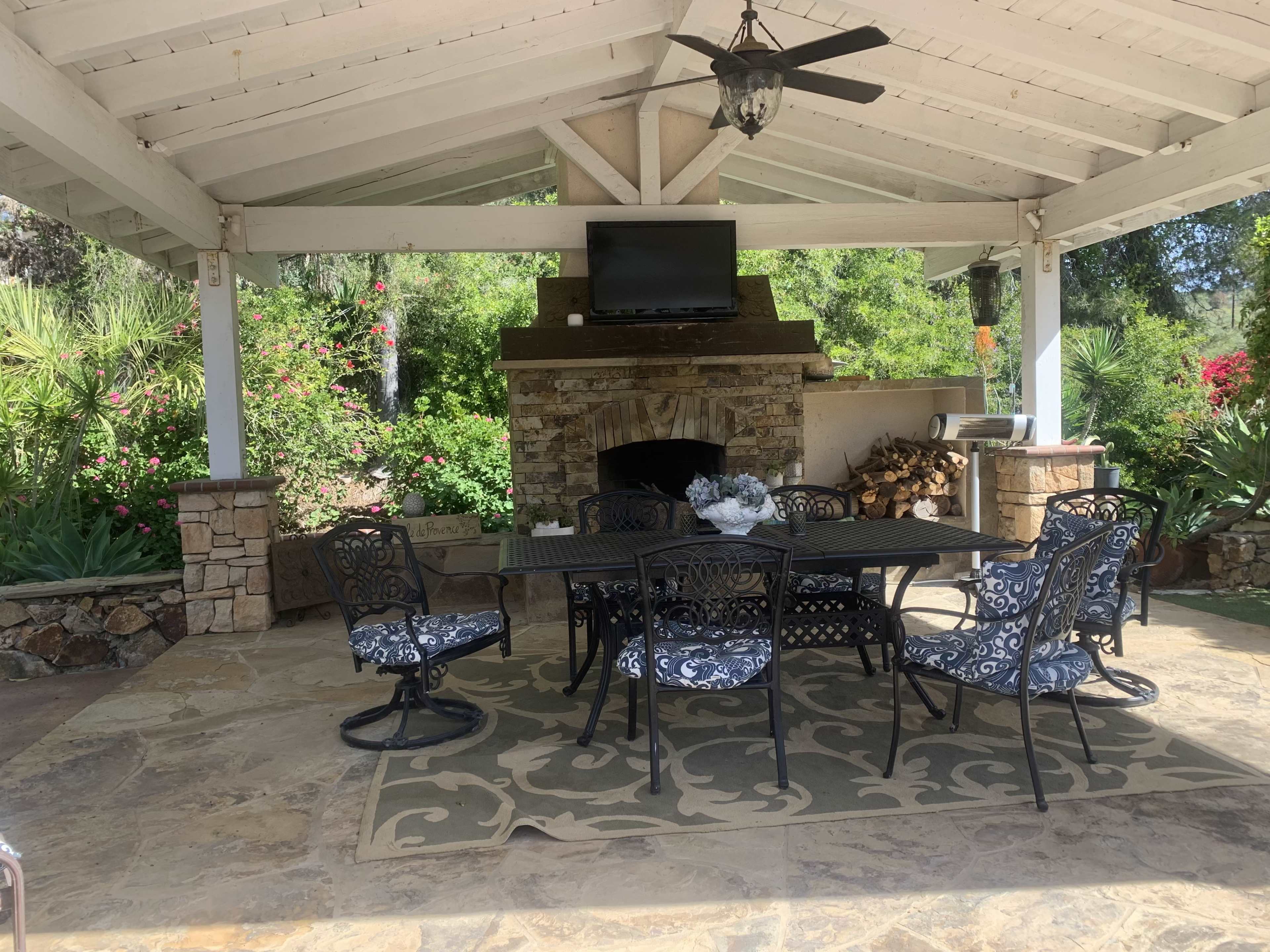 A covered outdoor patio features a large dining table surrounded by black metal chairs, with a stone fireplace and a television mounted on the wall.