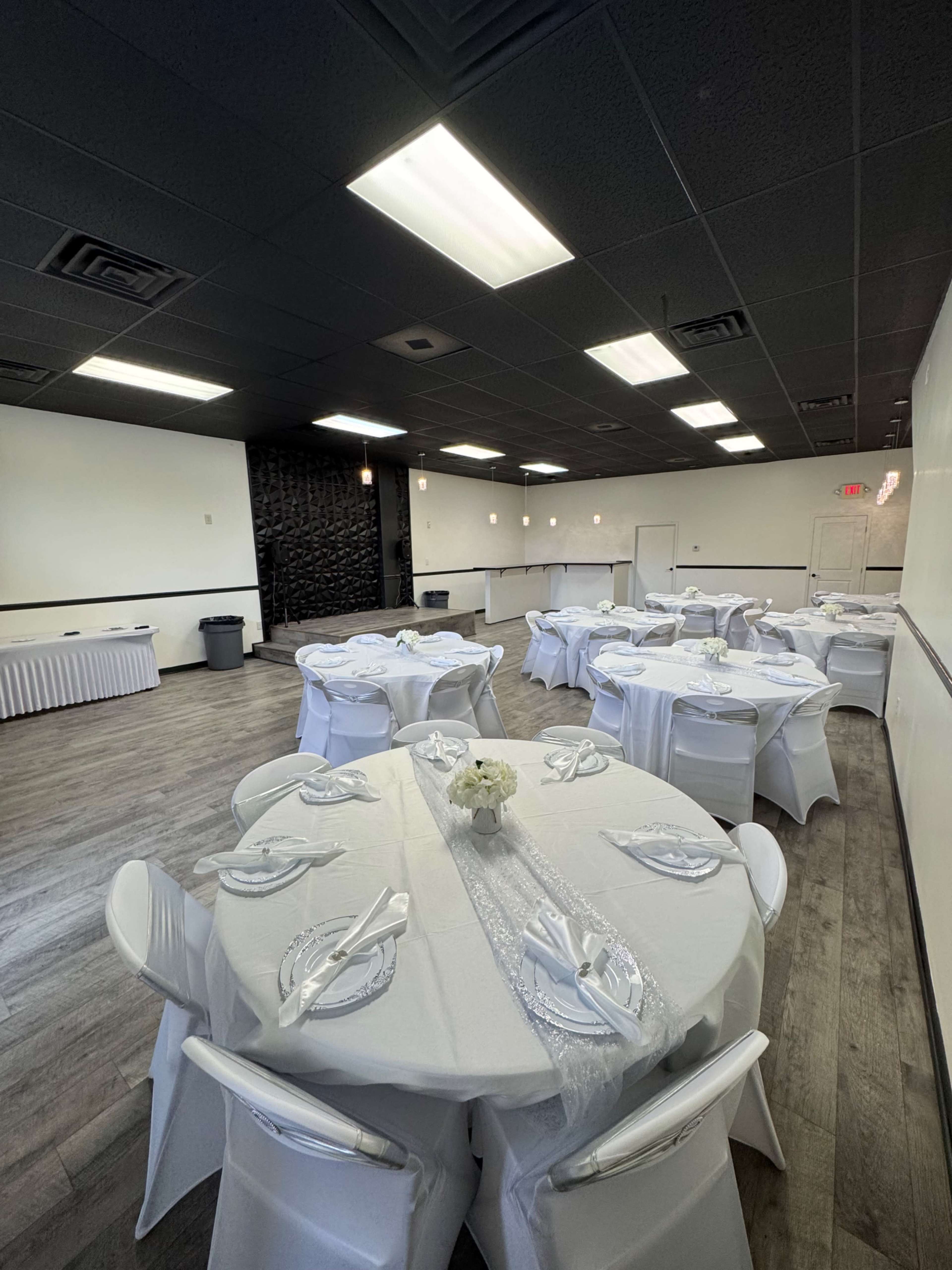 A spacious event room features several round tables set with white tablecloths and centerpieces, along with a decorative wall and overhead lighting.