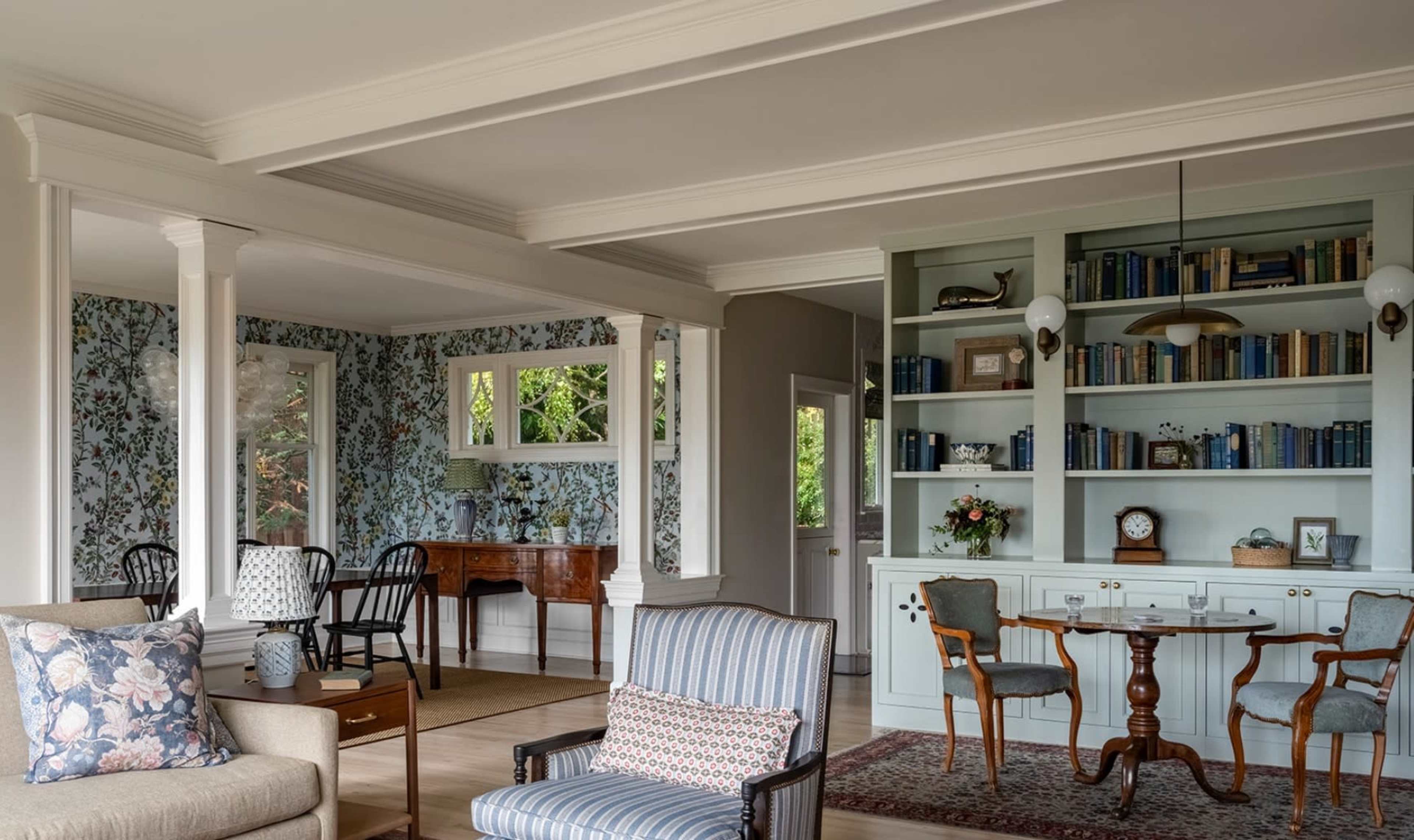 The image shows a well-lit living space featuring a mix of modern and classic furniture, with a bookshelf, floral wallpaper, and areas designated for seating and a small desk.