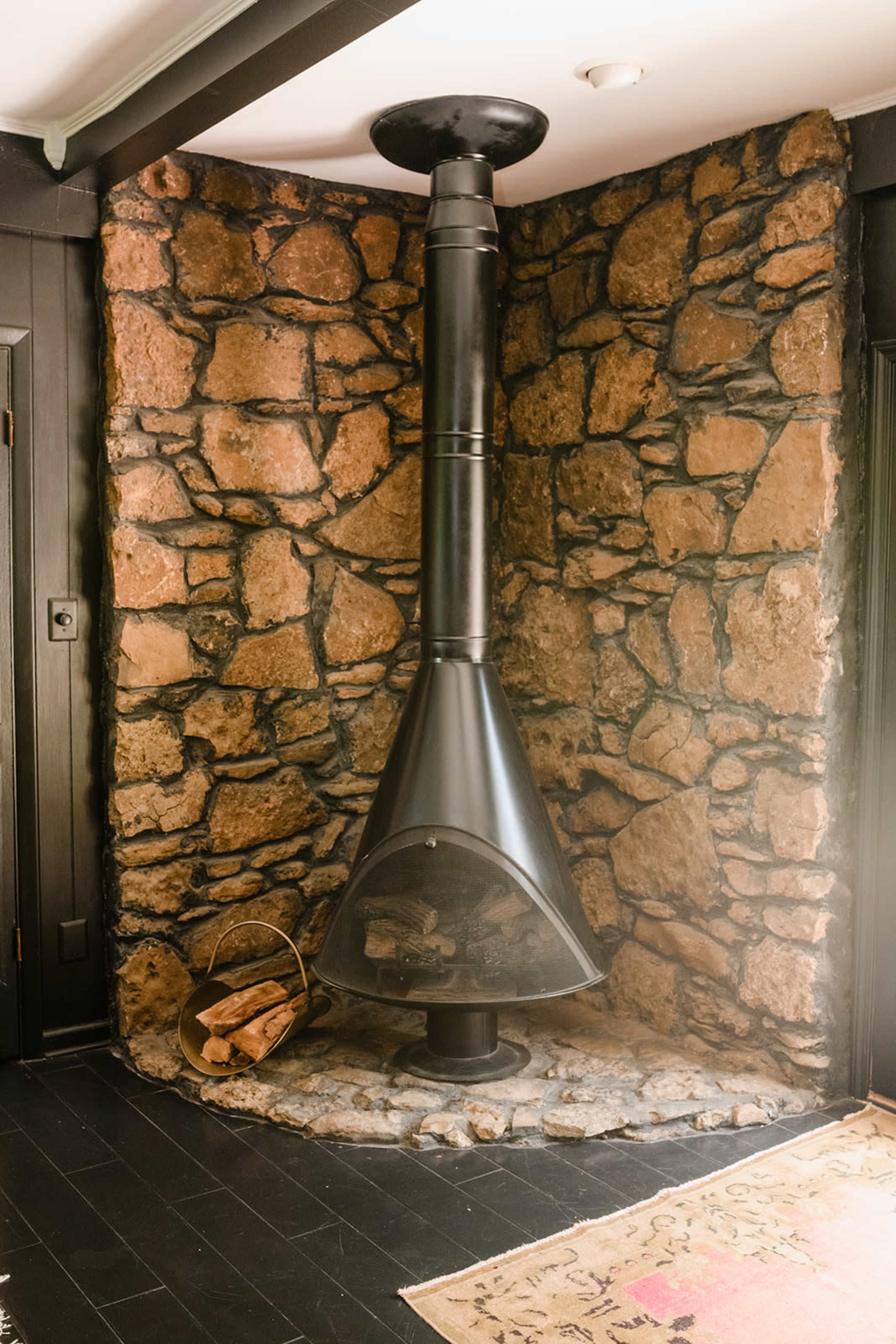 A black metal fireplace is installed between two stone walls in a corner of a room, with a basket of logs beside it.