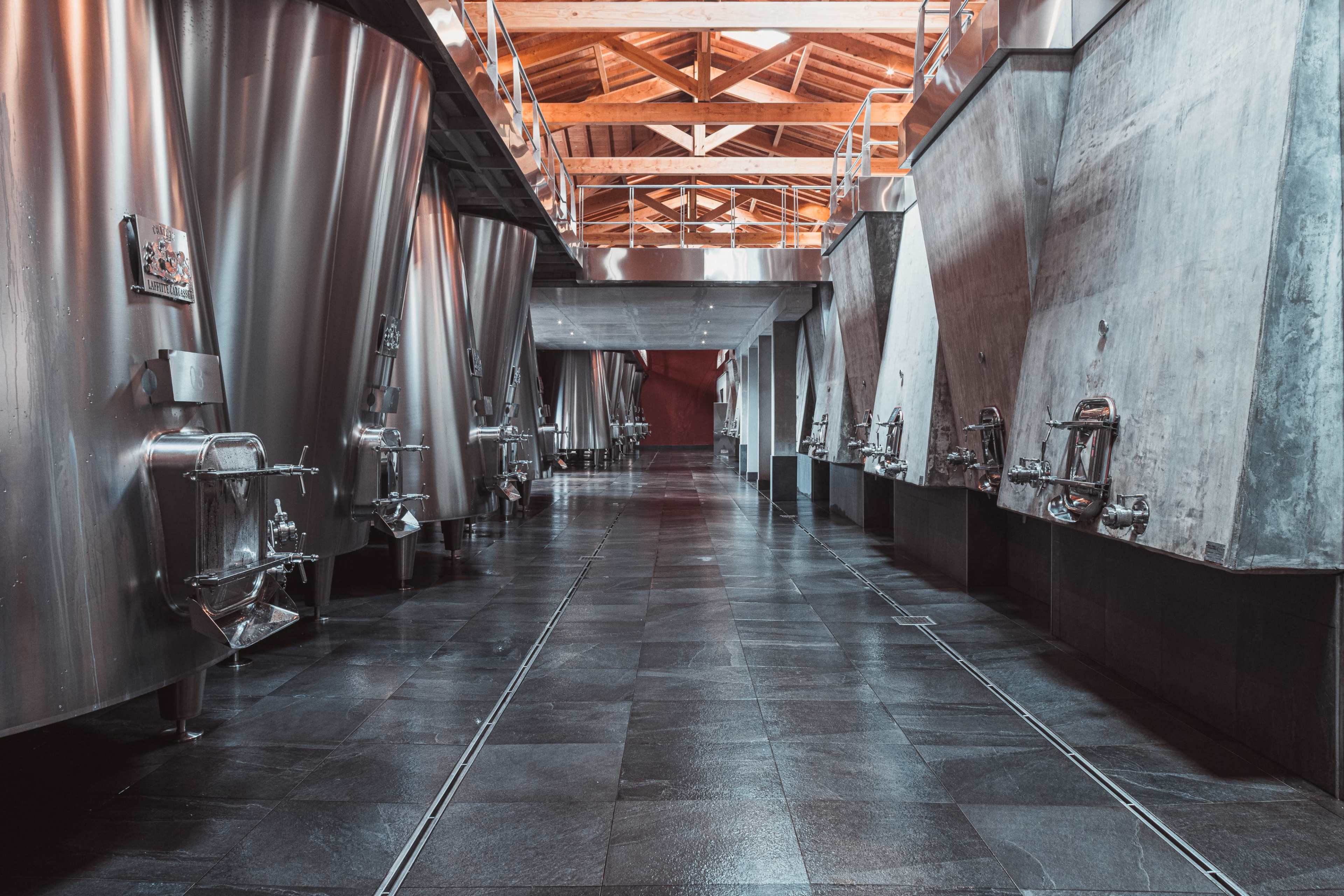 The image shows a long, industrial-style corridor lined with stainless steel fermentation tanks on both sides, under a wooden beam ceiling.