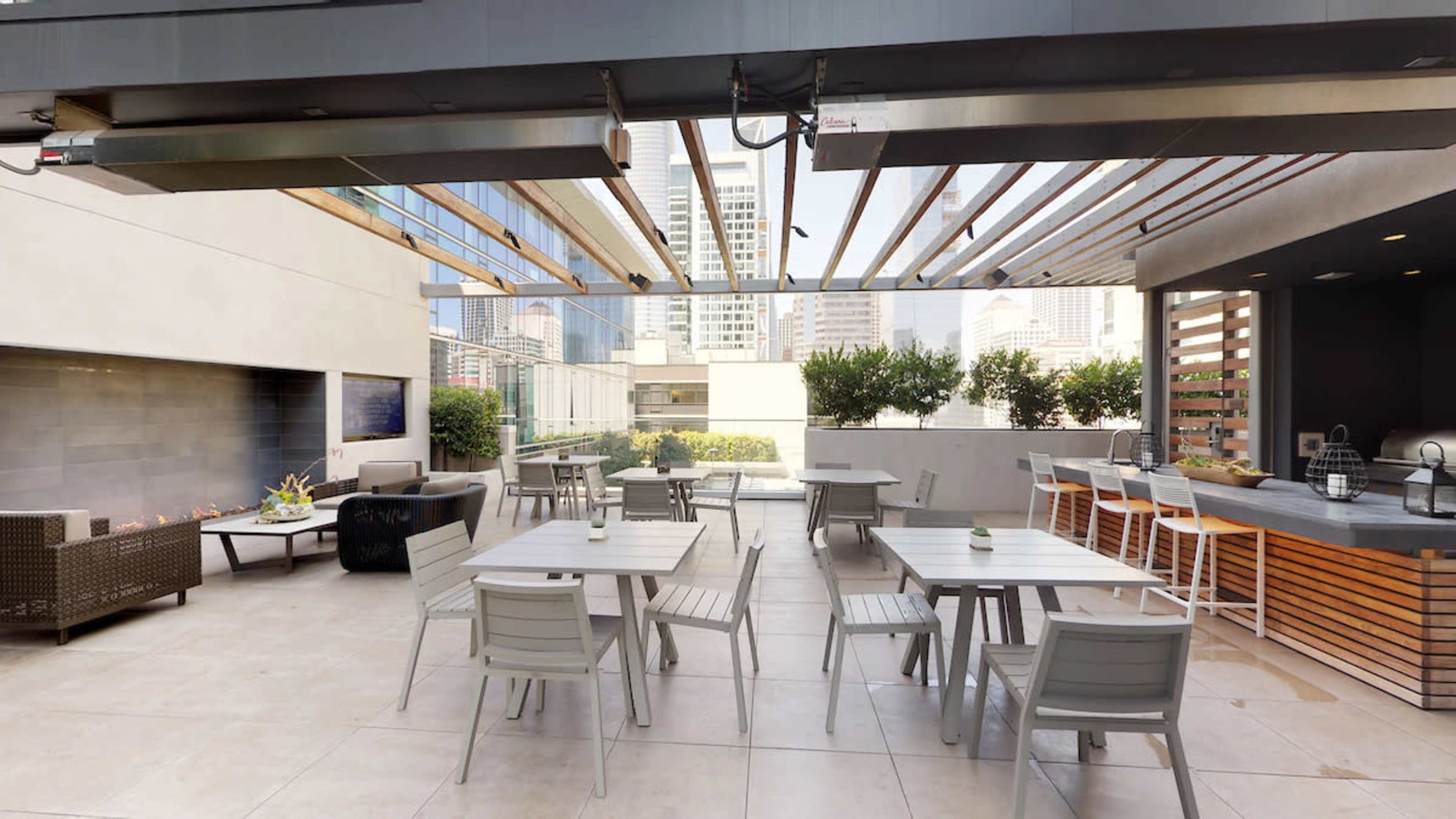 A modern outdoor seating area featuring tables and chairs under a slatted pergola, with greenery and a cooking area in the background.