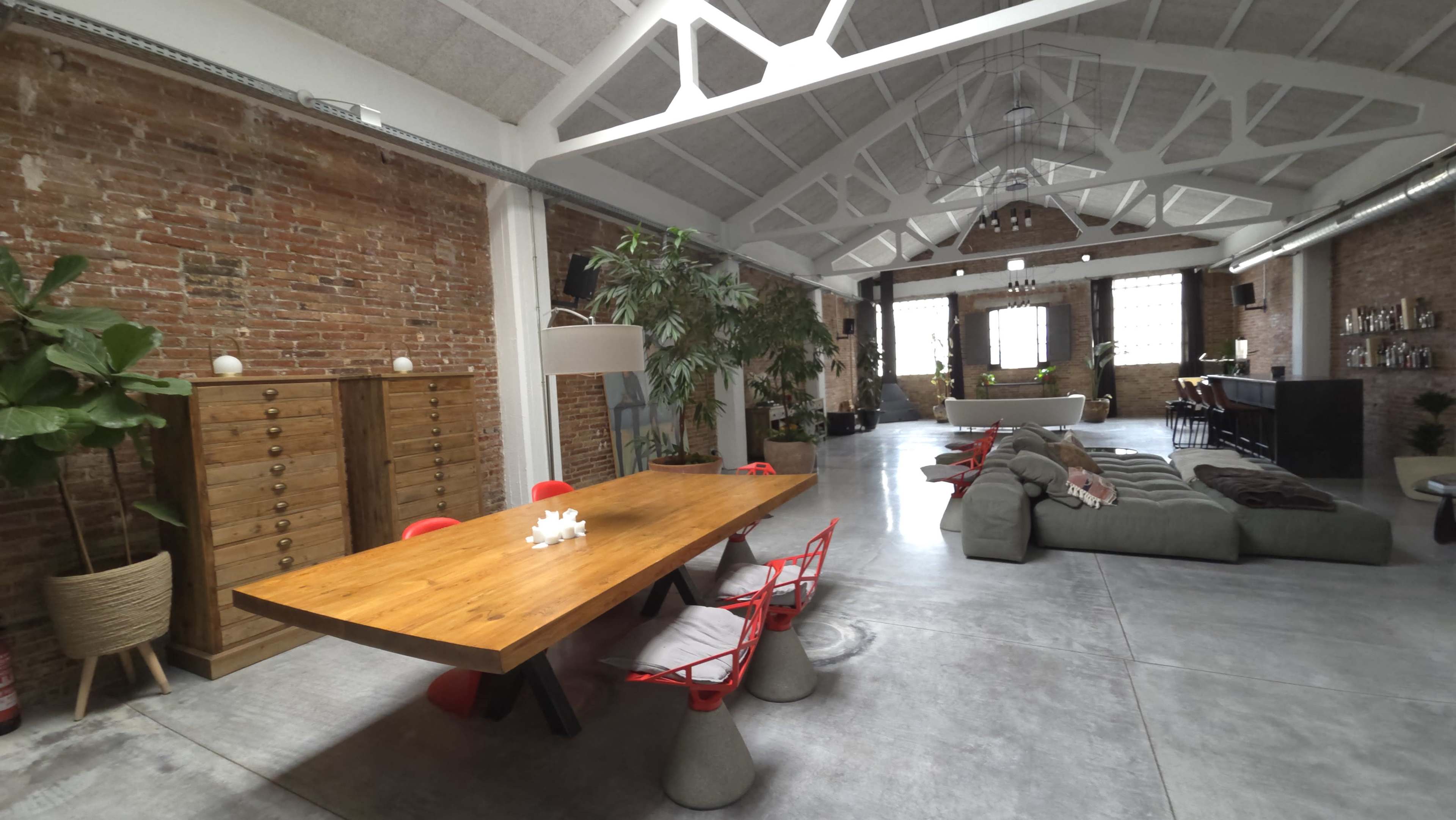 The image shows a spacious, industrial-style interior with a large wooden dining table surrounded by red chairs, complemented by a cozy seating area and exposed brick walls.