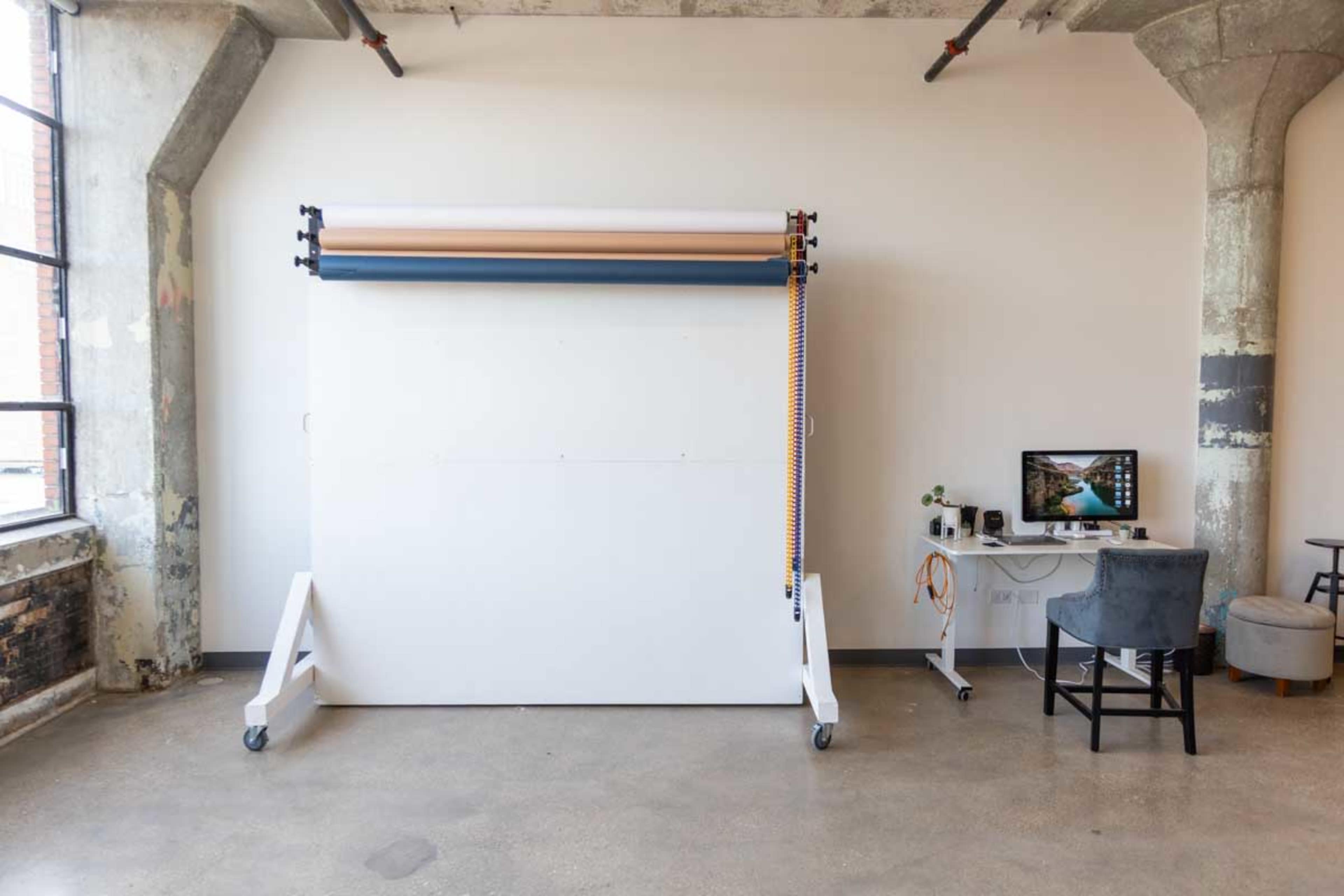 A large photography backdrop stand with rolls of paper is set against a bare wall in a spacious room, accompanied by a small desk and chair.