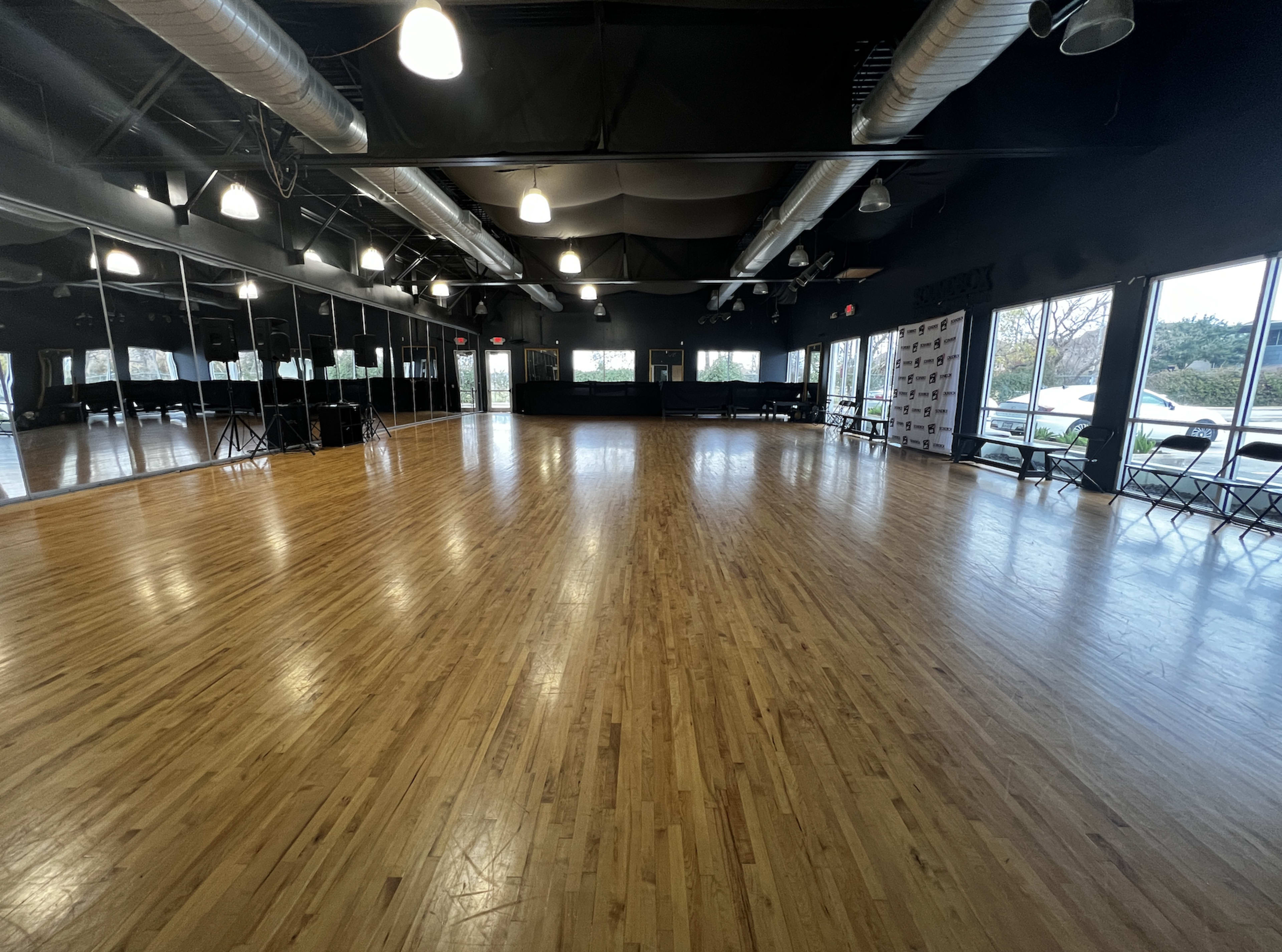 A spacious dance studio with hardwood flooring, large mirrors on the walls, and several empty chairs arranged along one side.