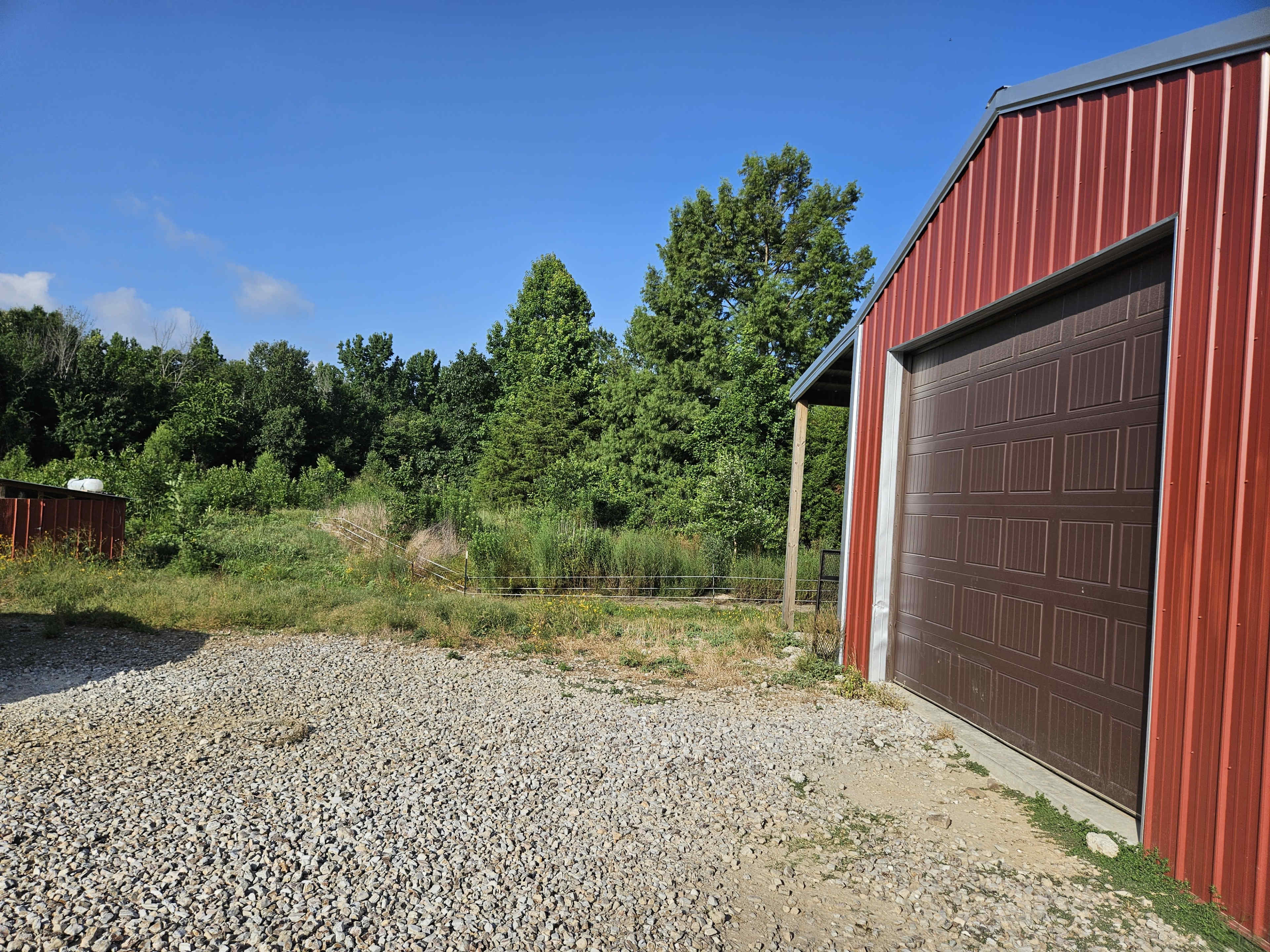 A red metal barn stands next to a gravel area with greenery in the background under a clear blue sky.