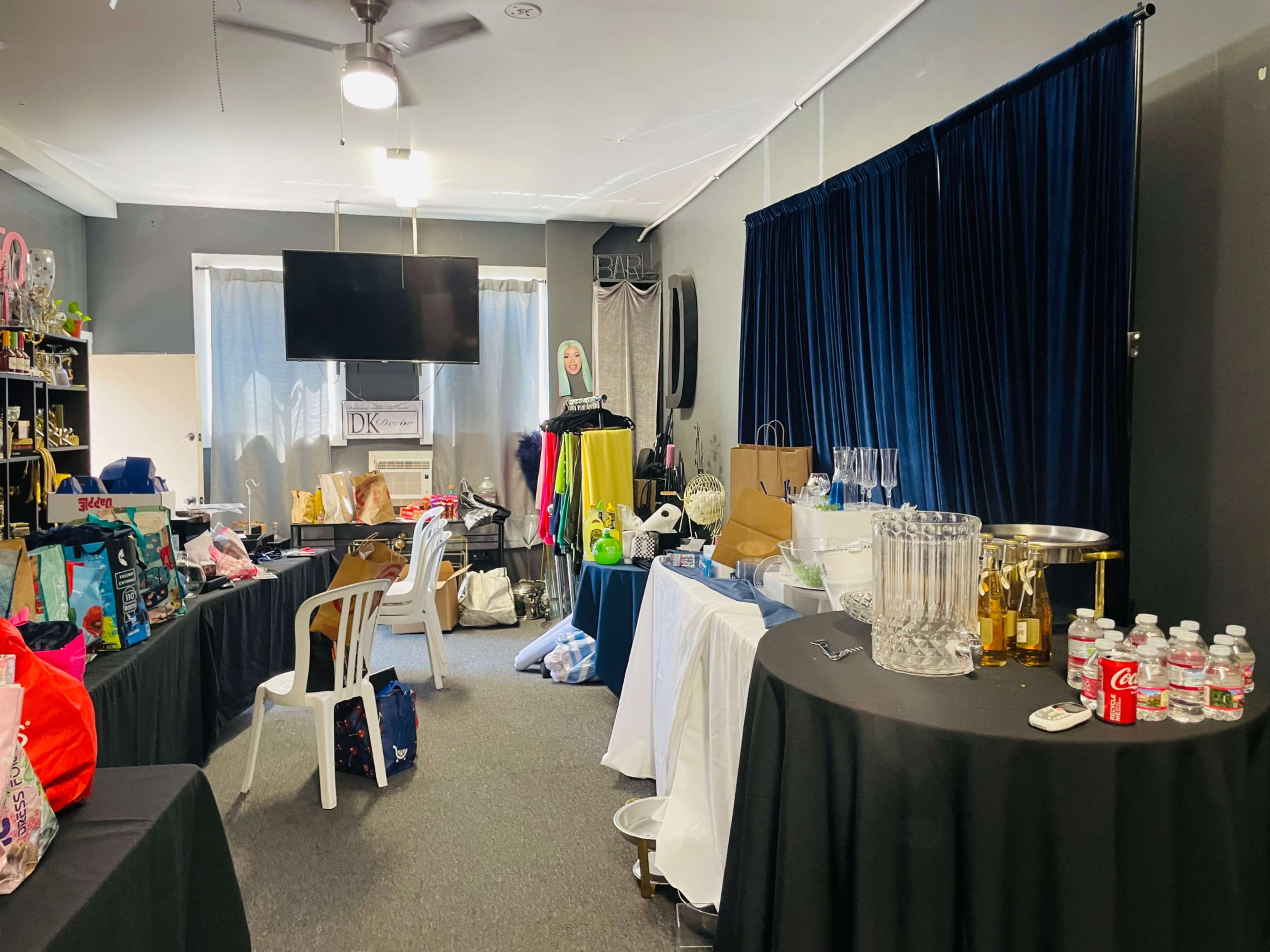 The image shows a cluttered room with multiple tables featuring drinks, bags, and various items, alongside a backdrop and a large television.