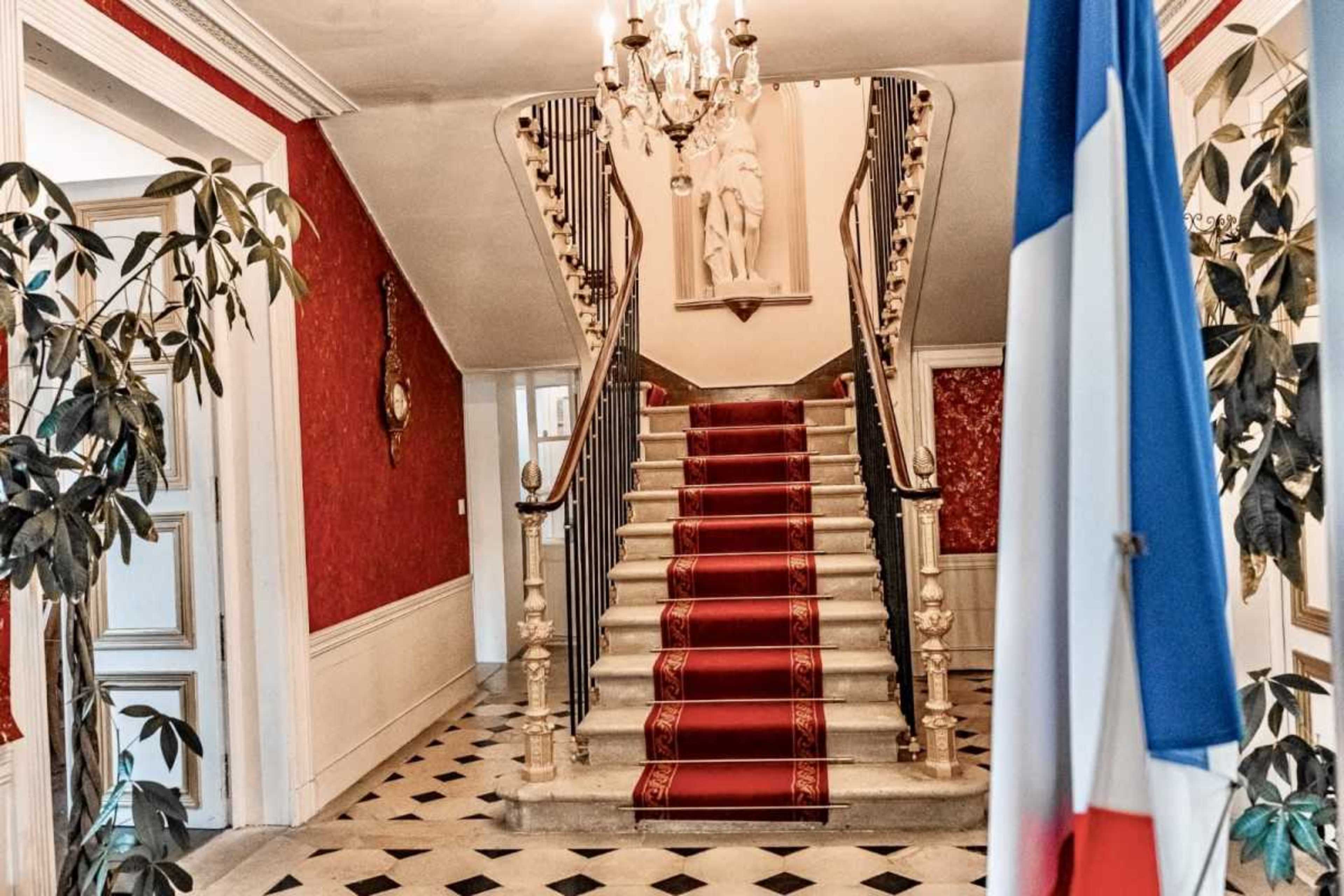 A grand staircase with a red carpet leads up to a landing, flanked by indoor plants and a French flag in the foreground.