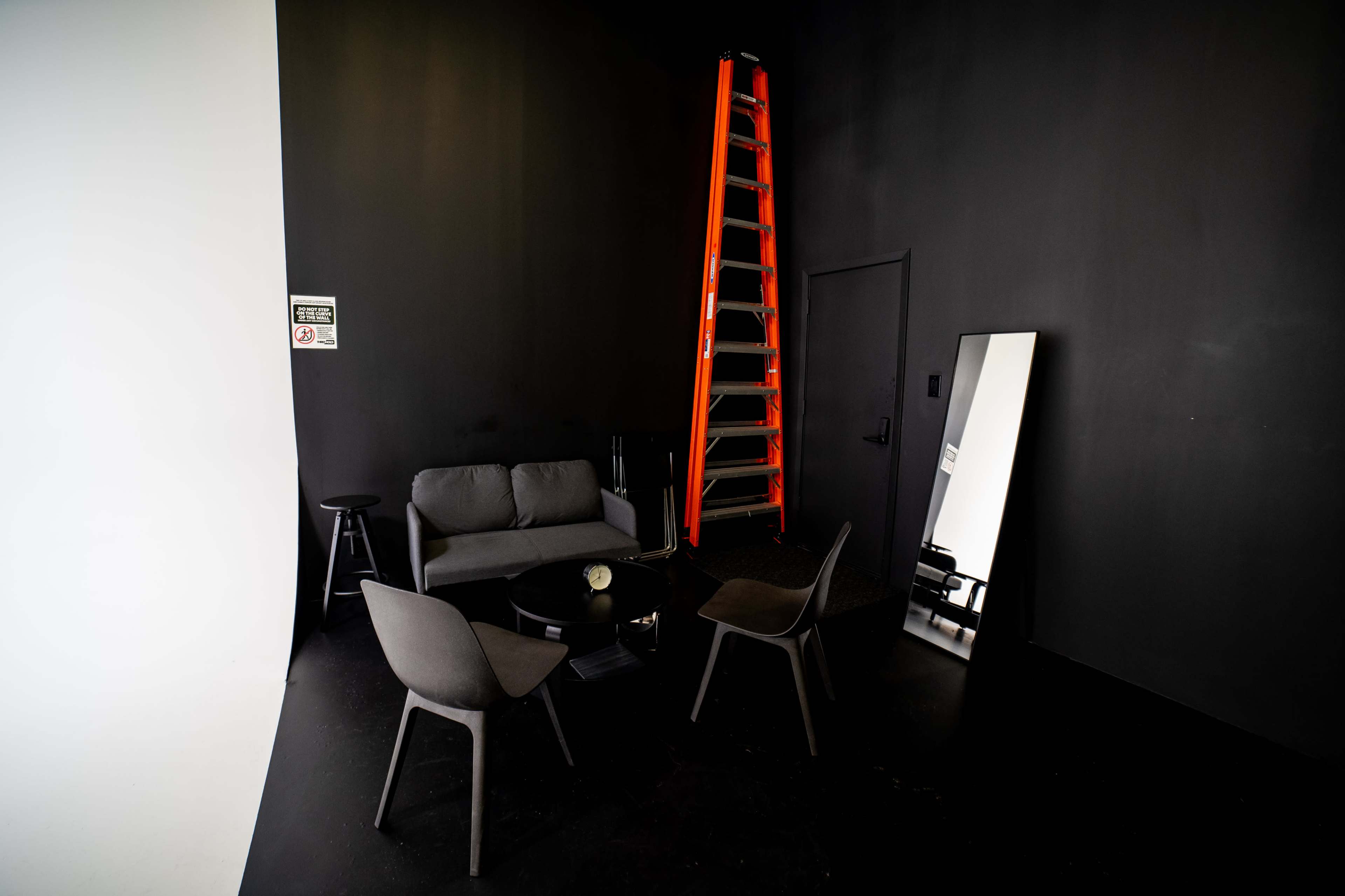 A small, dimly lit room features a gray sofa, a round table, four chairs, a tall ladder against a dark wall, and a large mirror.