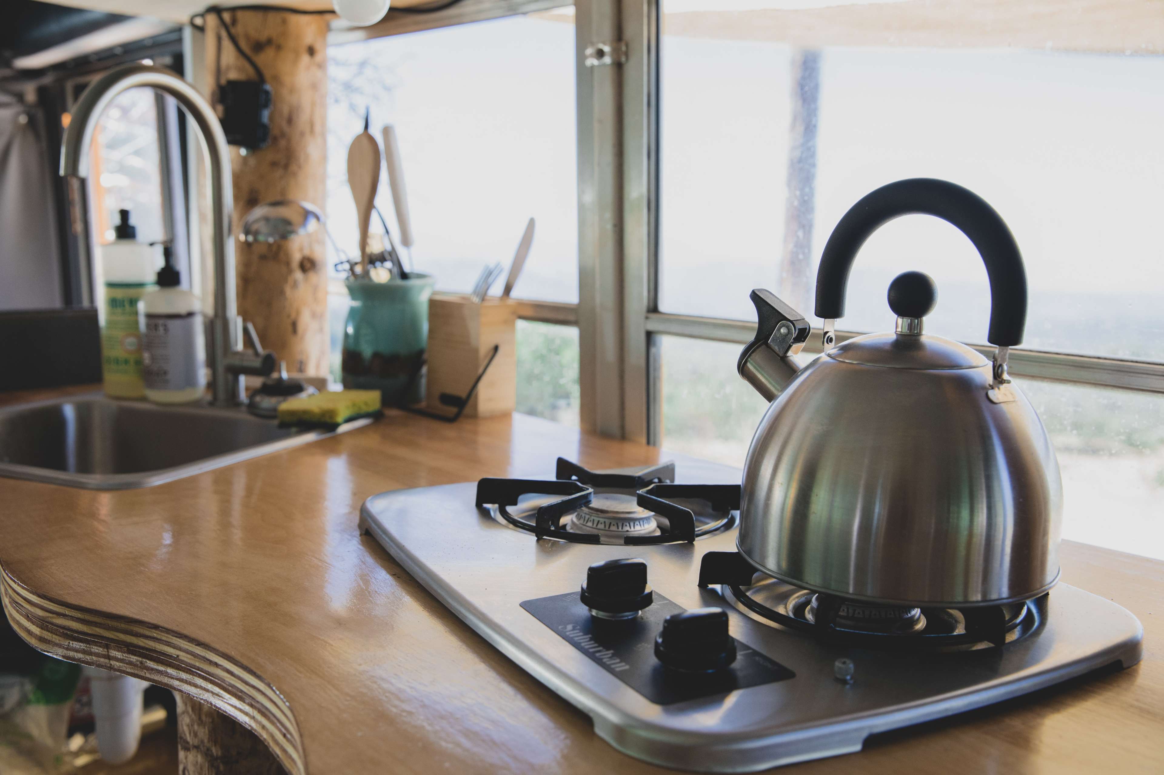 A stainless steel kettle sits on a gas stovetop in a kitchen with a wooden countertop and a window view.