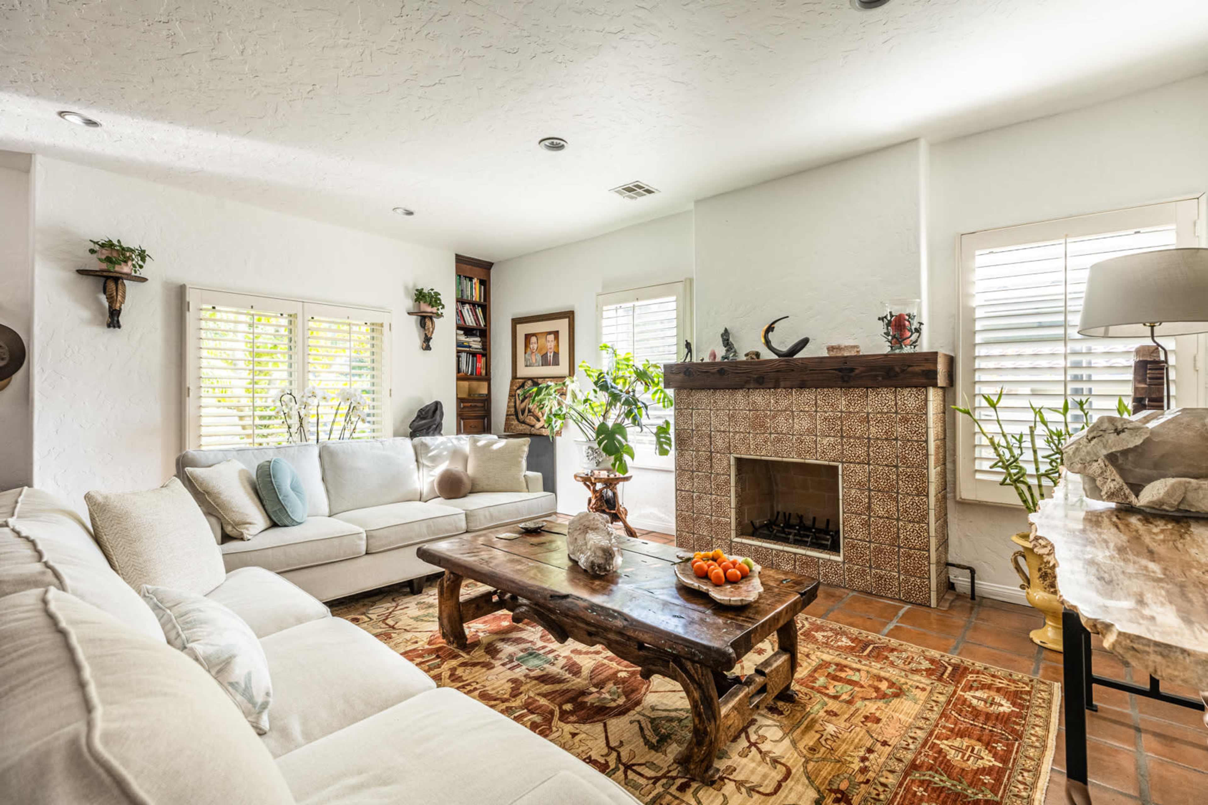 A living room features a white couch, a wooden coffee table with a bowl of fruit, and a tiled fireplace, surrounded by plants and decorative items.