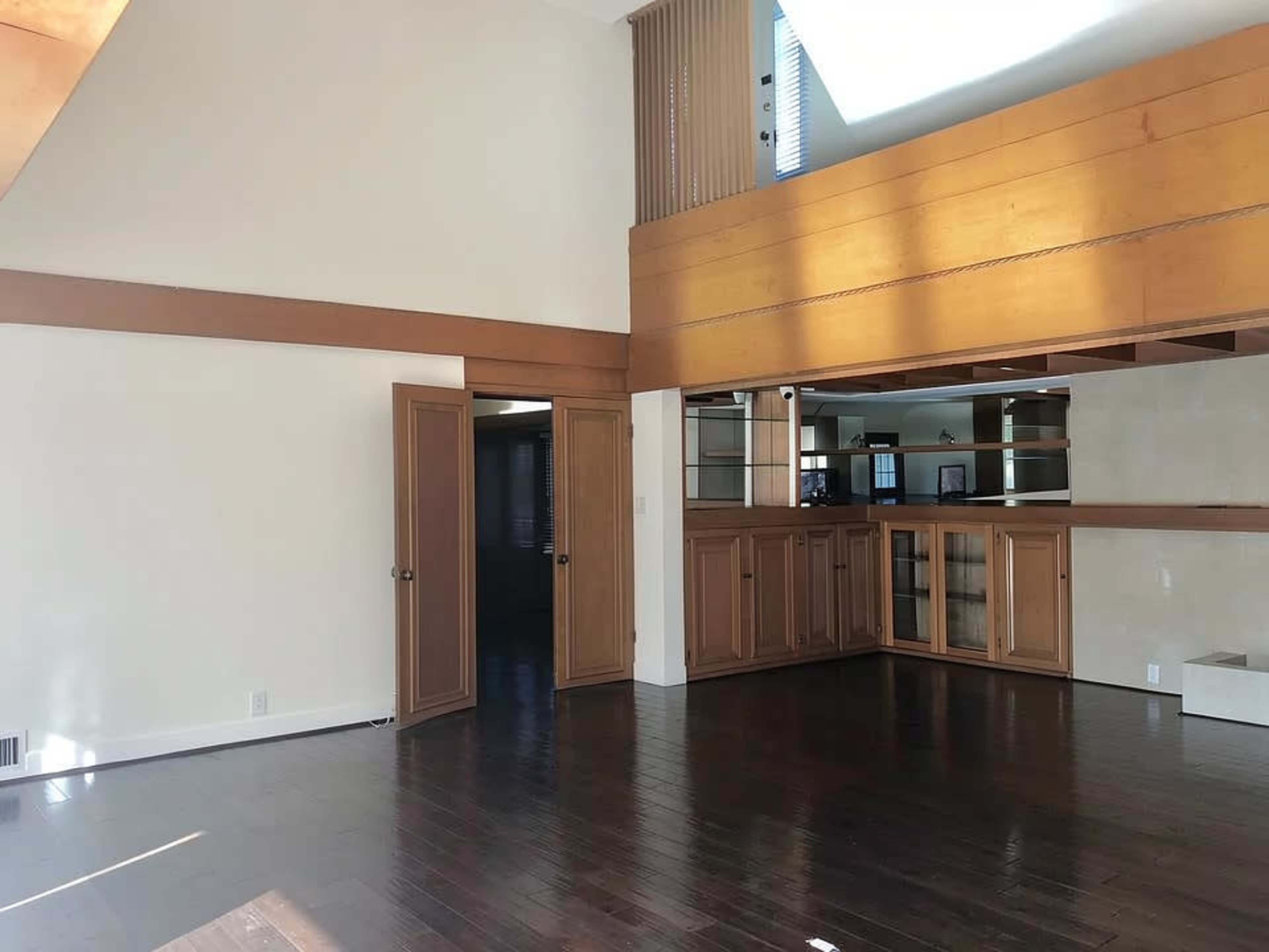 The image shows a spacious living area with wooden flooring, a high ceiling, and a section with glass shelving and cabinetry.