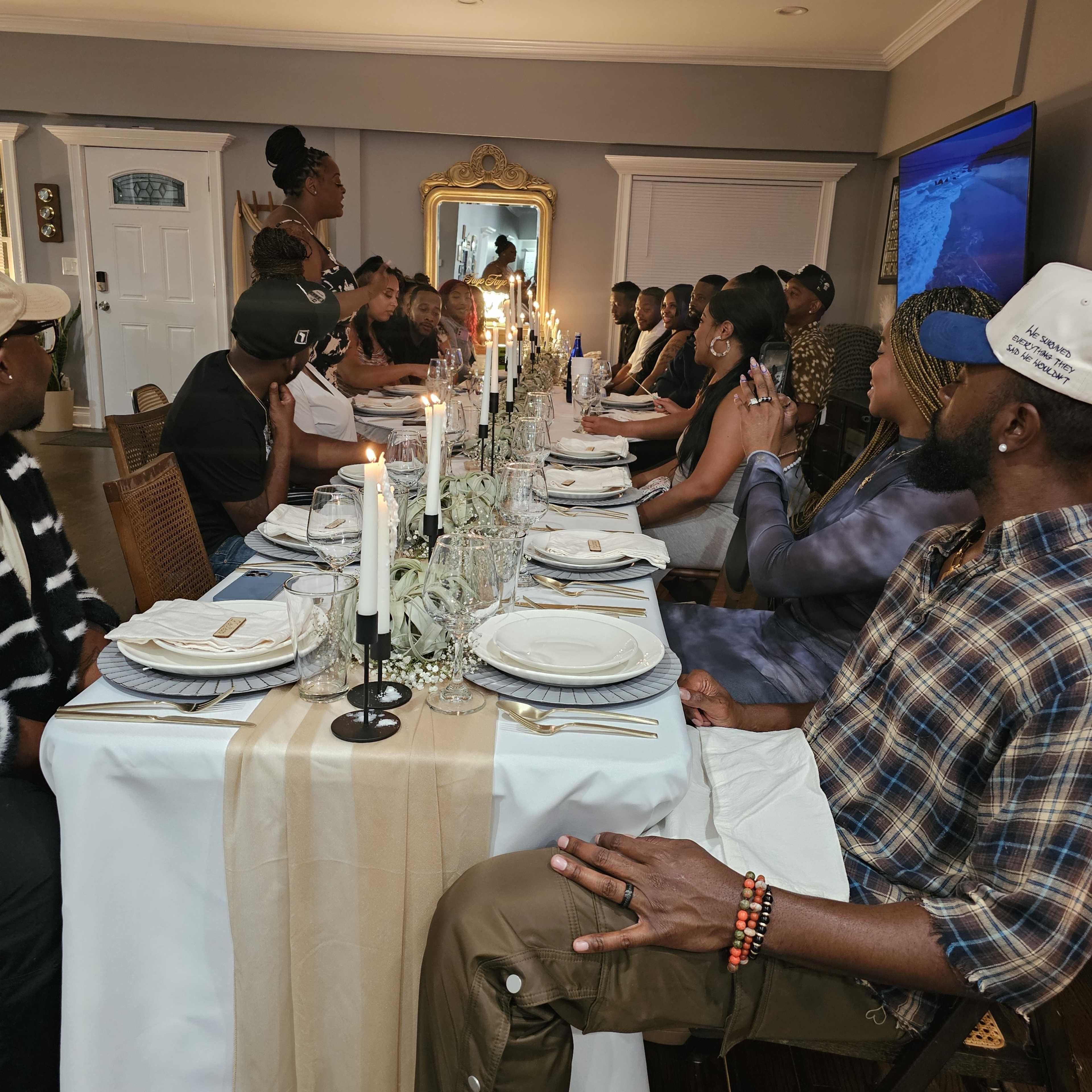 A long dining table is set with plates and candles, populated by guests engaged in conversation during a gathering in a well-decorated room.
