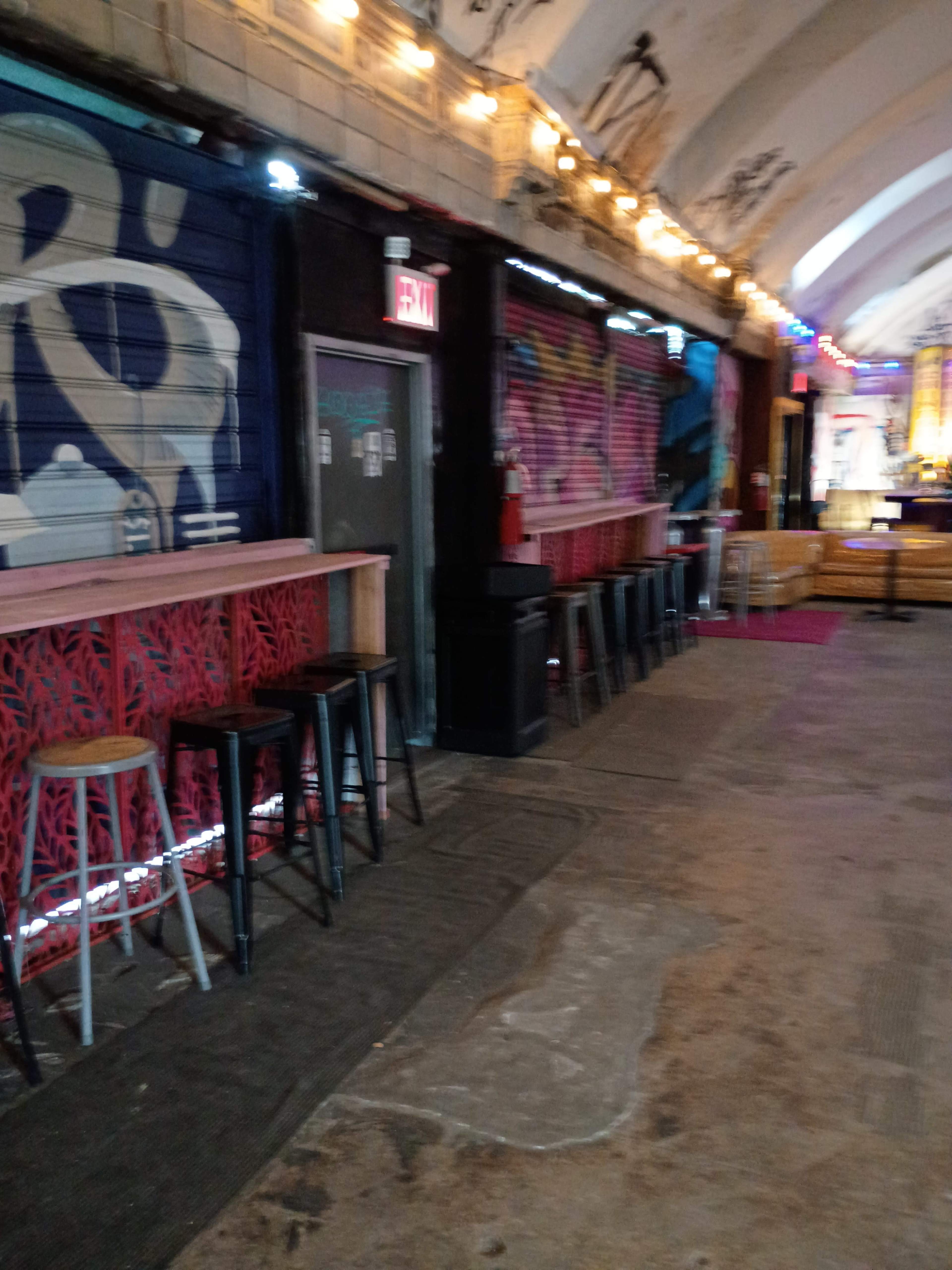 The image shows a narrow, colorful indoor space with a bar area, featuring stools along a mural-adorned wall and string lights overhead.