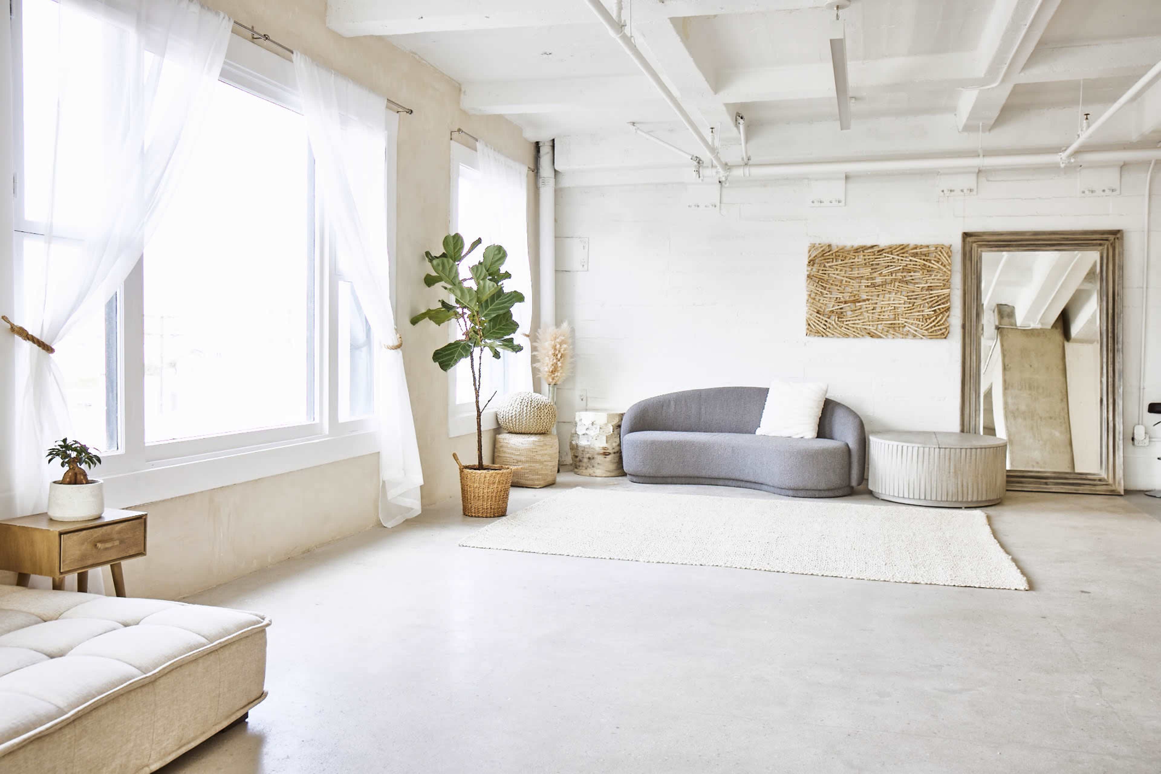 A bright, minimalist living space features large windows, a gray curved sofa, a woven wall hanging, and plants in decorative pots.