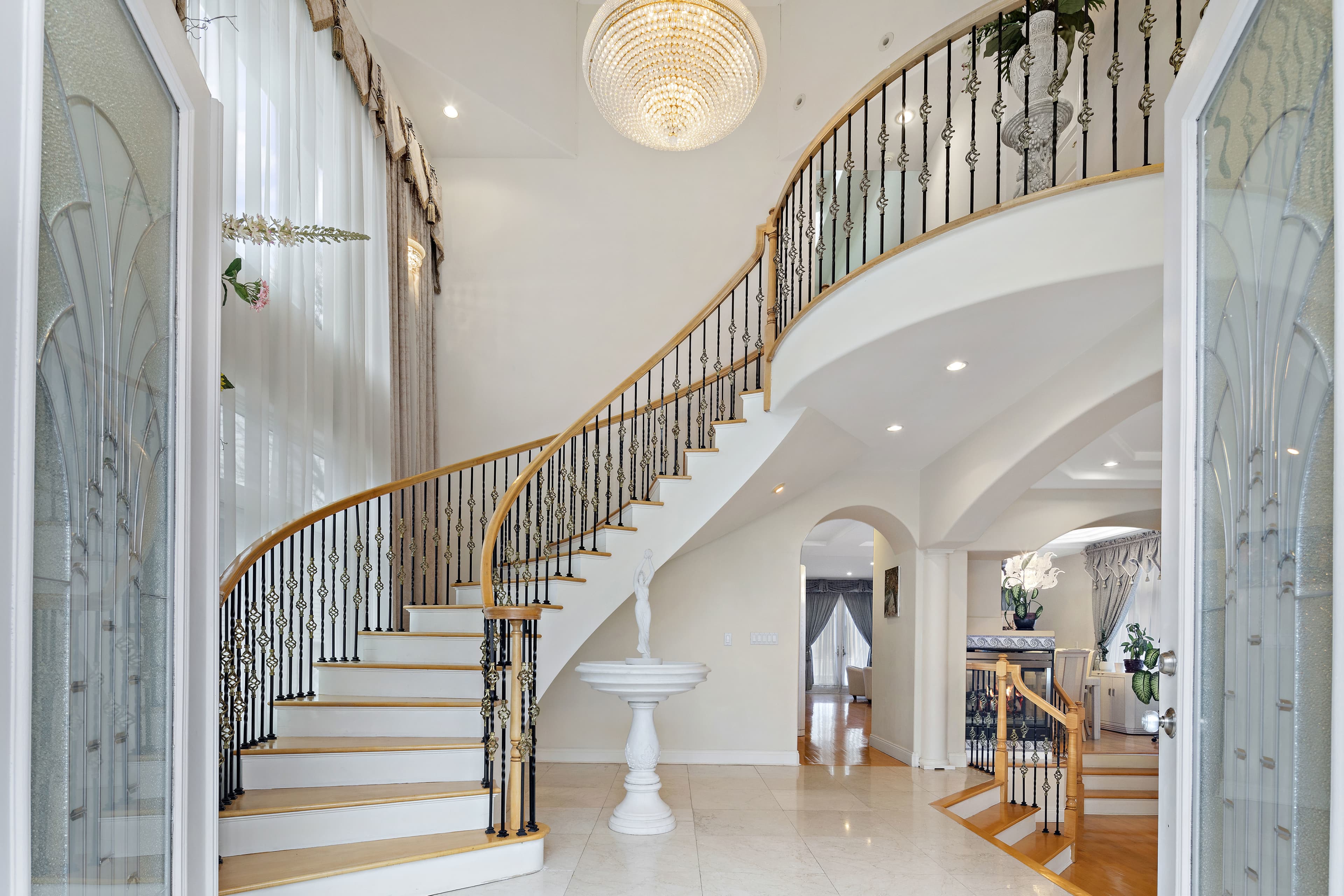 A grand foyer with a curved staircase, polished marble flooring, and a large chandelier overhead.