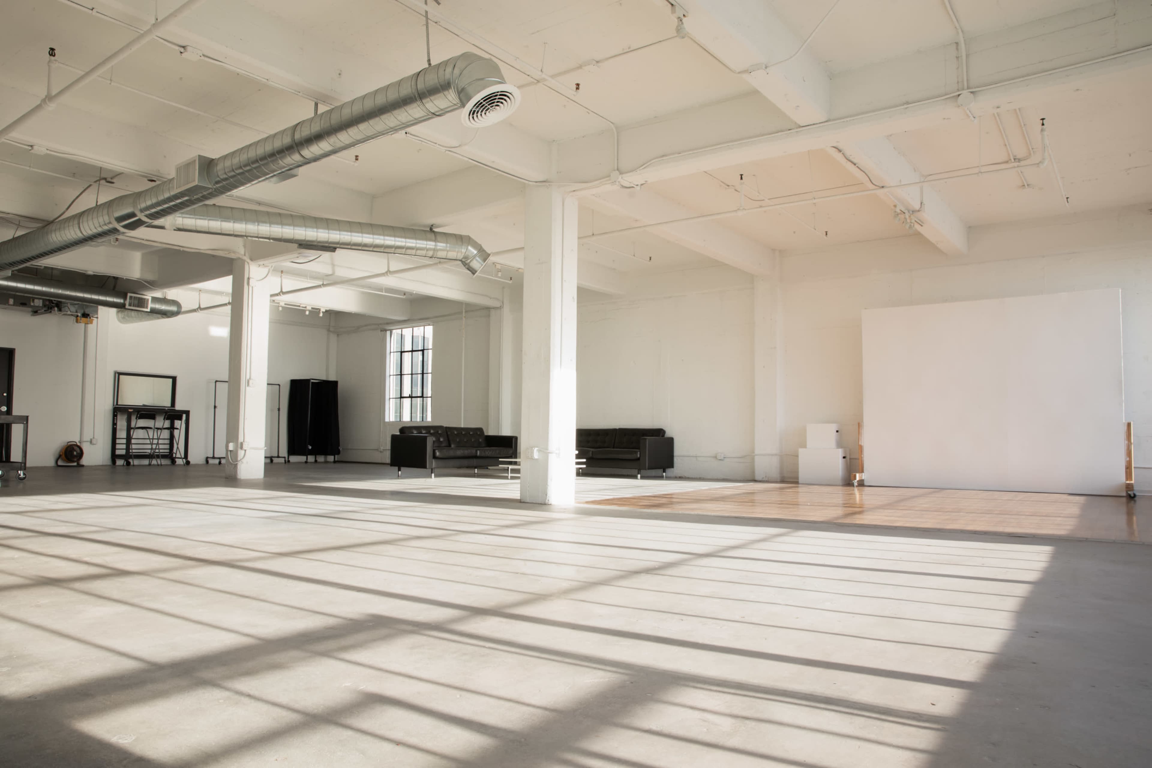 The image shows a spacious, sunlit studio with concrete flooring, large windows, and minimal furnishings including a black couch and a white backdrop.