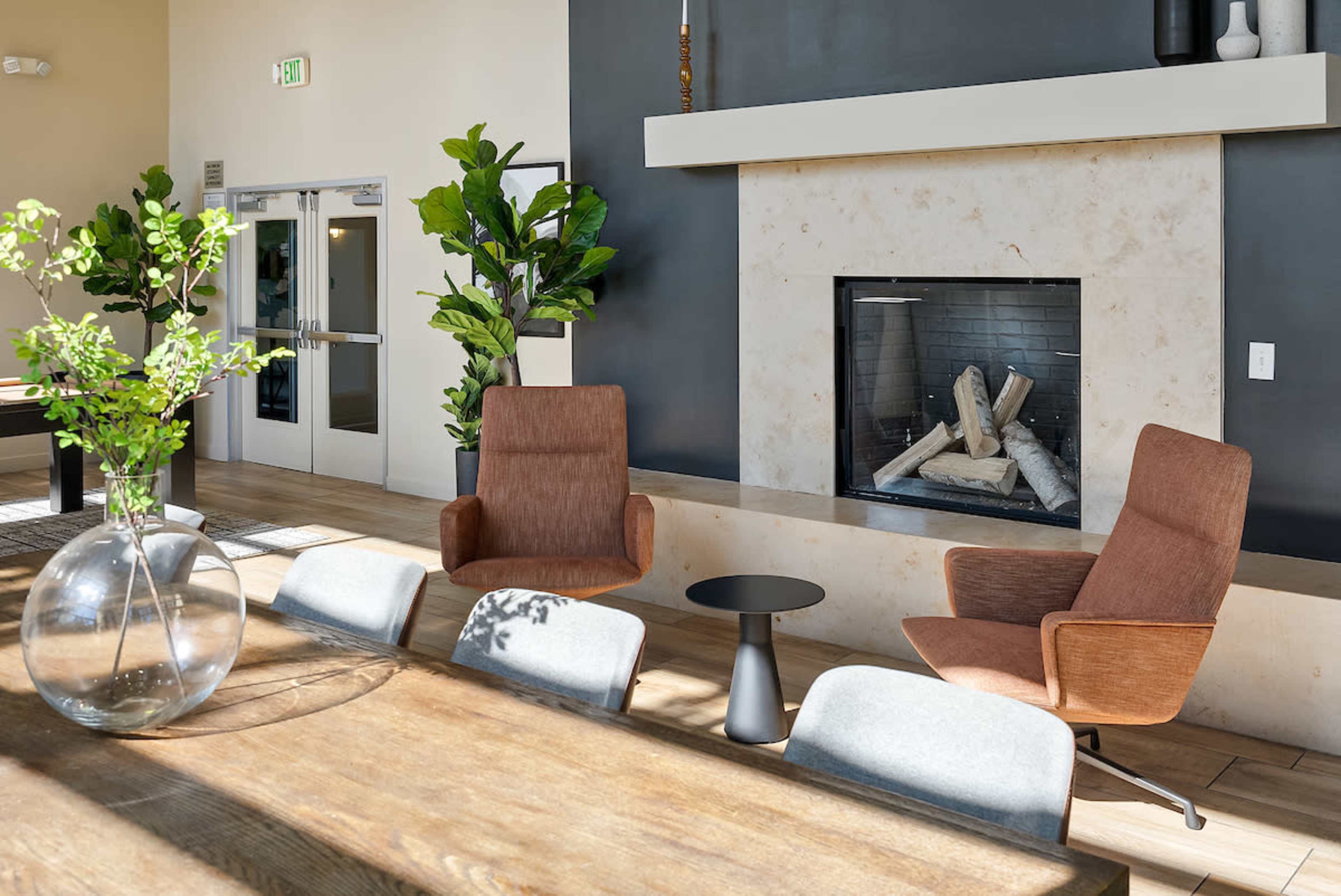 The image shows a modern interior space featuring a dining table, two armchairs, a small side table, and a fireplace surrounded by plants.