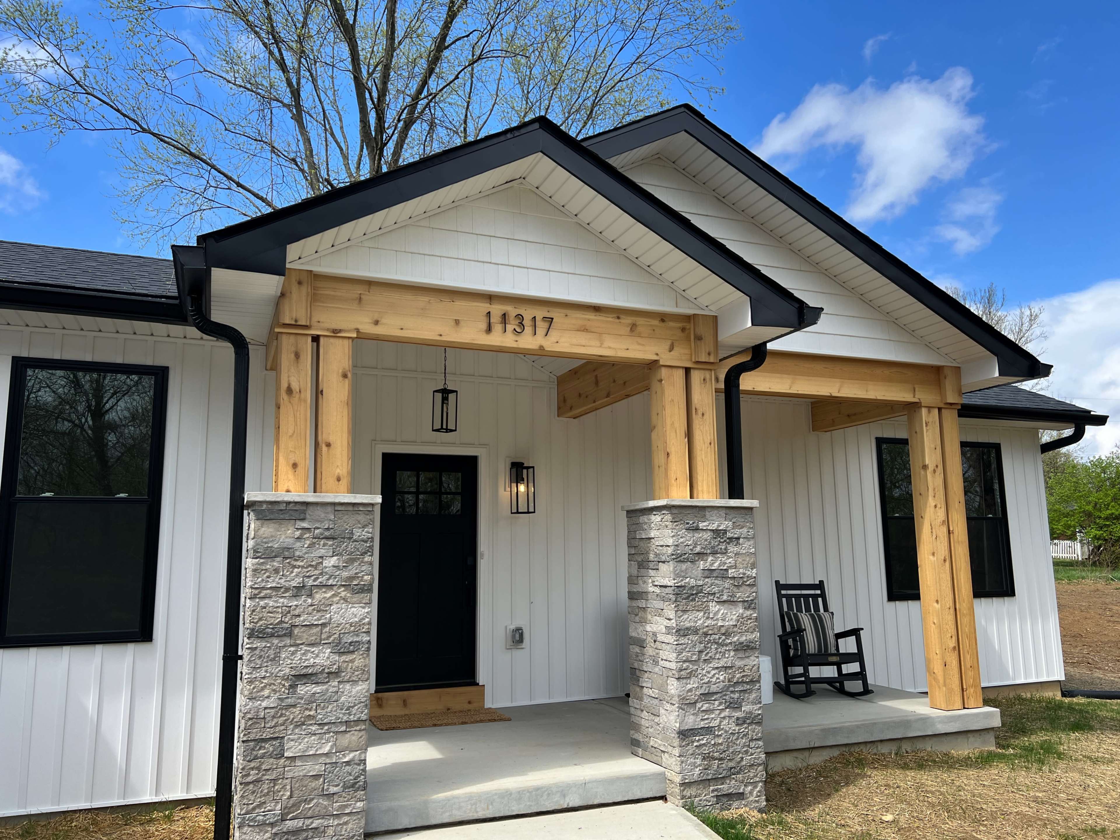 The image shows a modern-style house with a white exterior, featuring wooden beams over the entrance and a stone base, along with a black front door and a rocking chair on the porch.