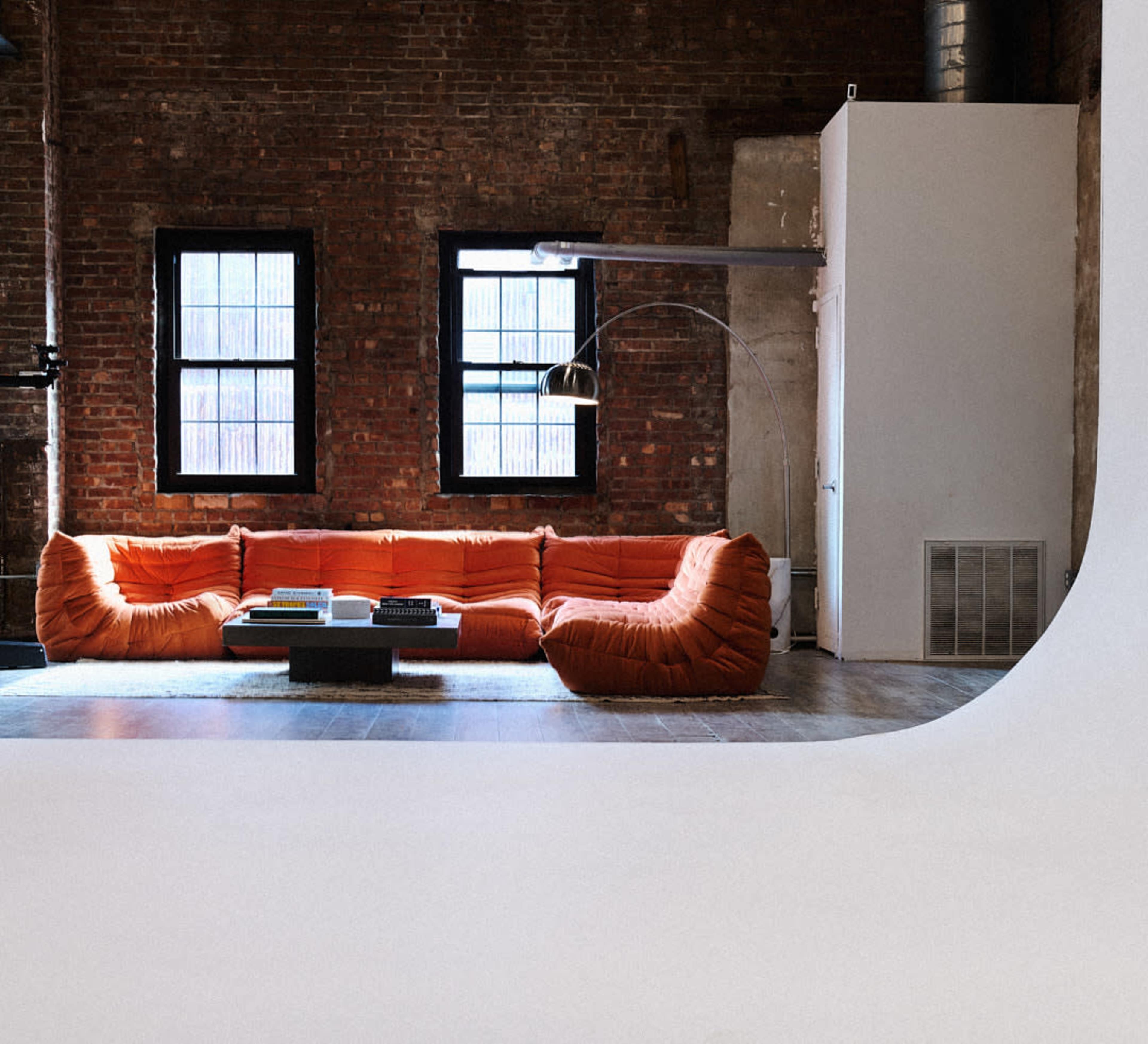 A modern living room with a bright orange sectional sofa, two windows revealing a brick wall, and a minimalist coffee table.