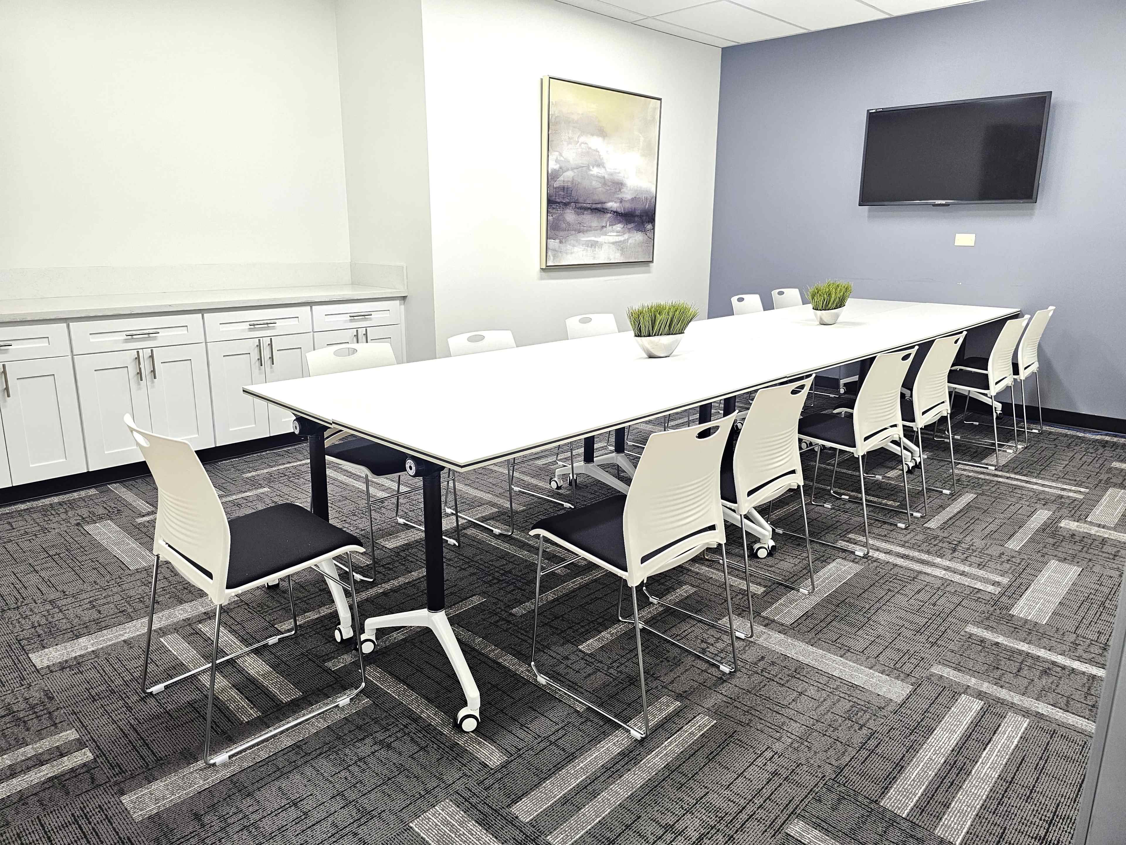 A long, white conference table is set up in a meeting room surrounded by twelve chairs, with a flat-screen TV on the wall and a side cabinet in the background.