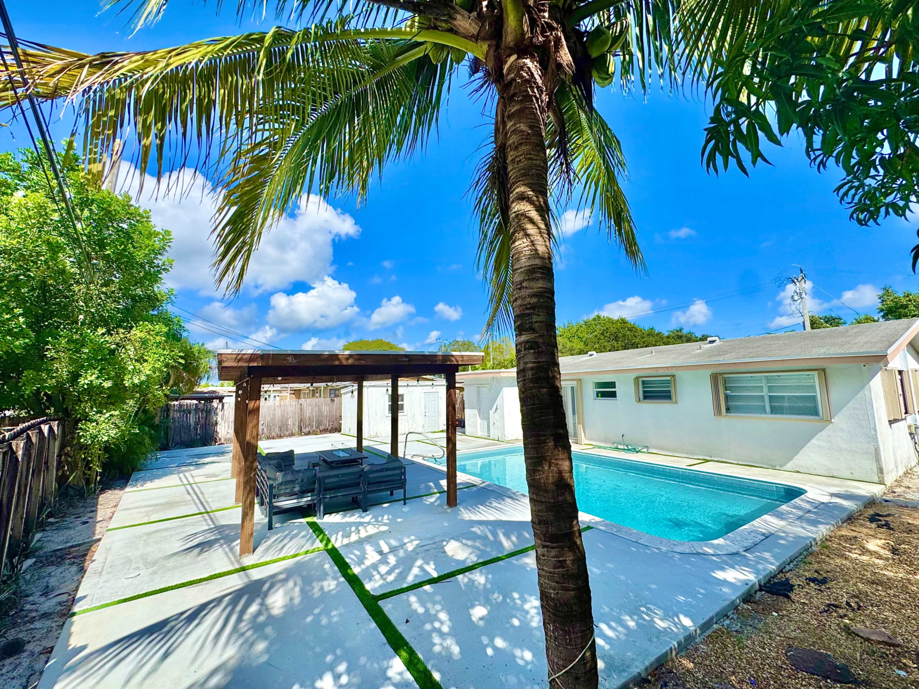 The image shows a backyard with a swimming pool, a wooden pergola, and a palm tree, surrounded by green foliage and a clear blue sky.