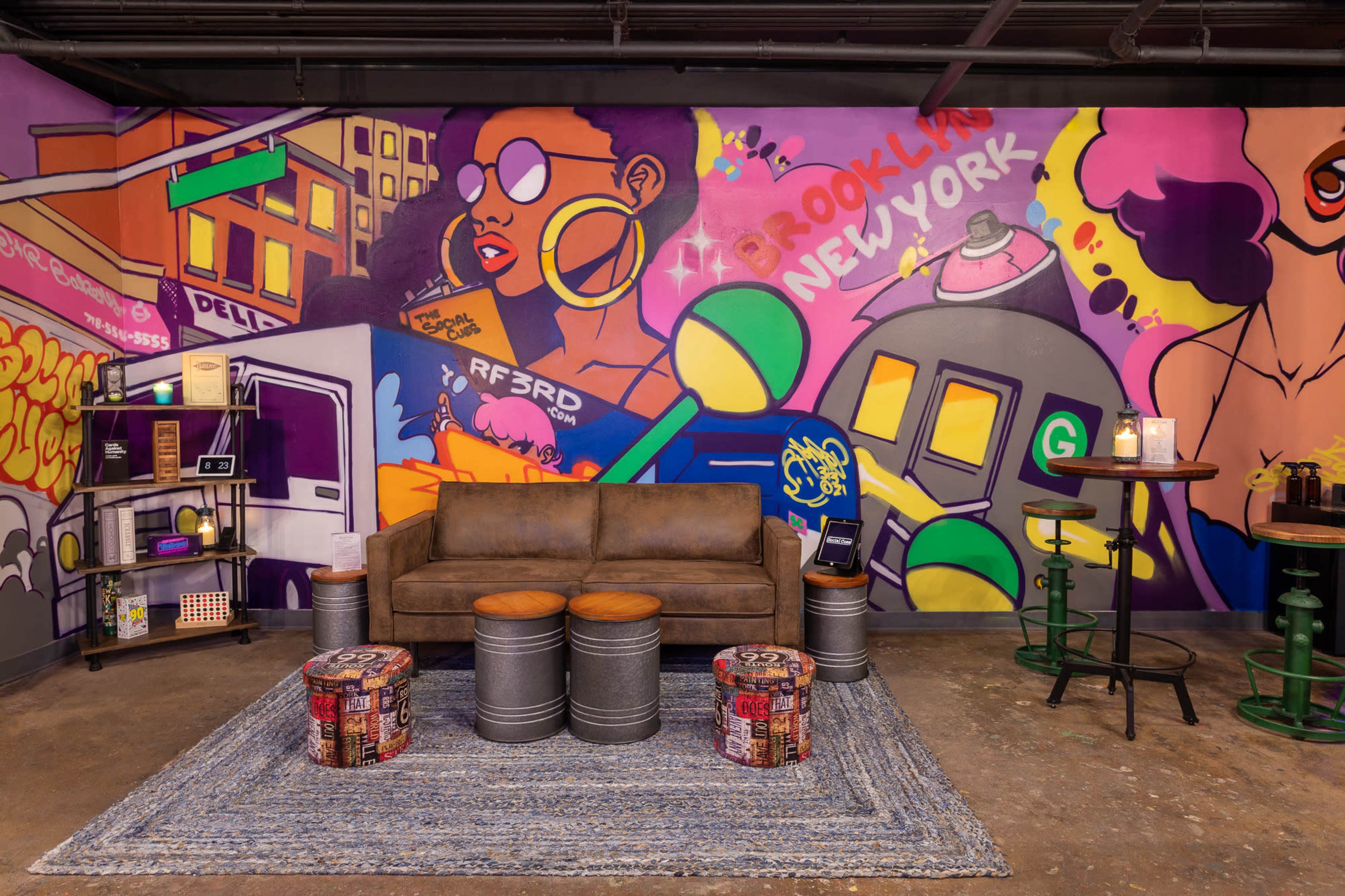 A modern urban space featuring a brown leather sofa, colorful graffiti art on the walls, and industrial-style furniture, including metal stools and a woven rug.