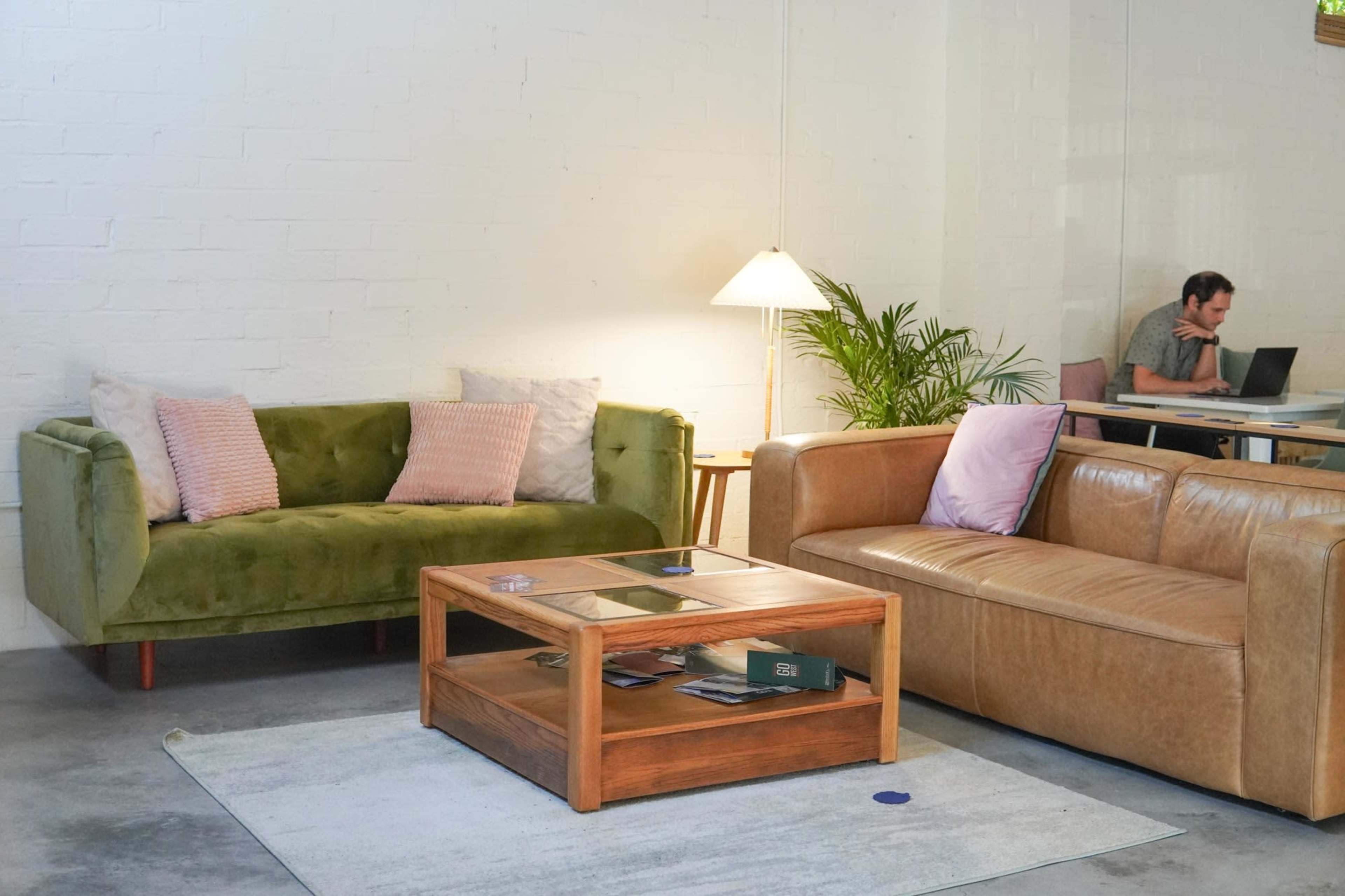 A green couch and a brown leather sofa are situated in a bright room with a wooden coffee table and a lamp, while a person works on a laptop at a nearby table.