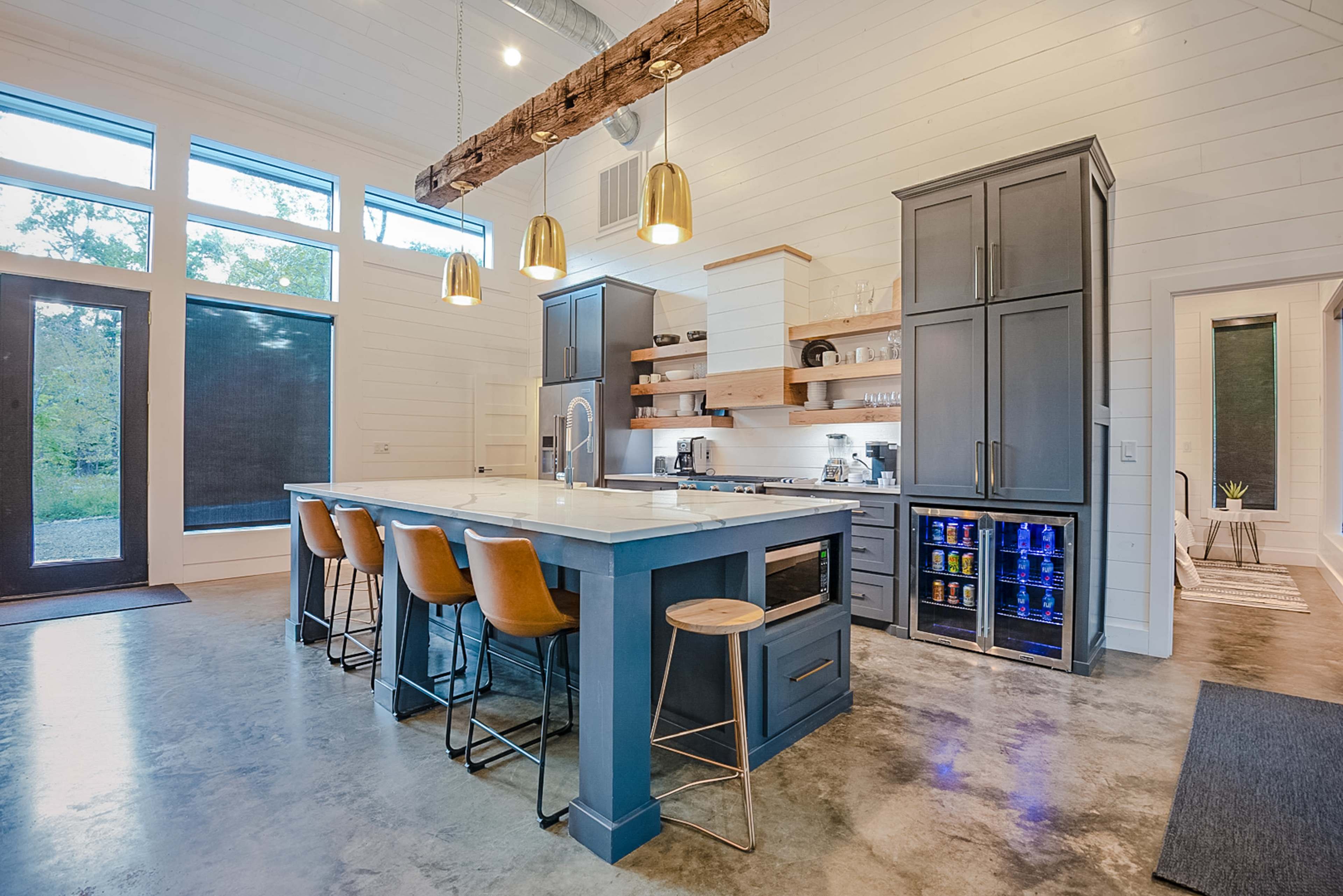 A modern kitchen with a large island, wooden bar stools, and a combination of gray cabinetry and white walls, illuminated by pendant lights.