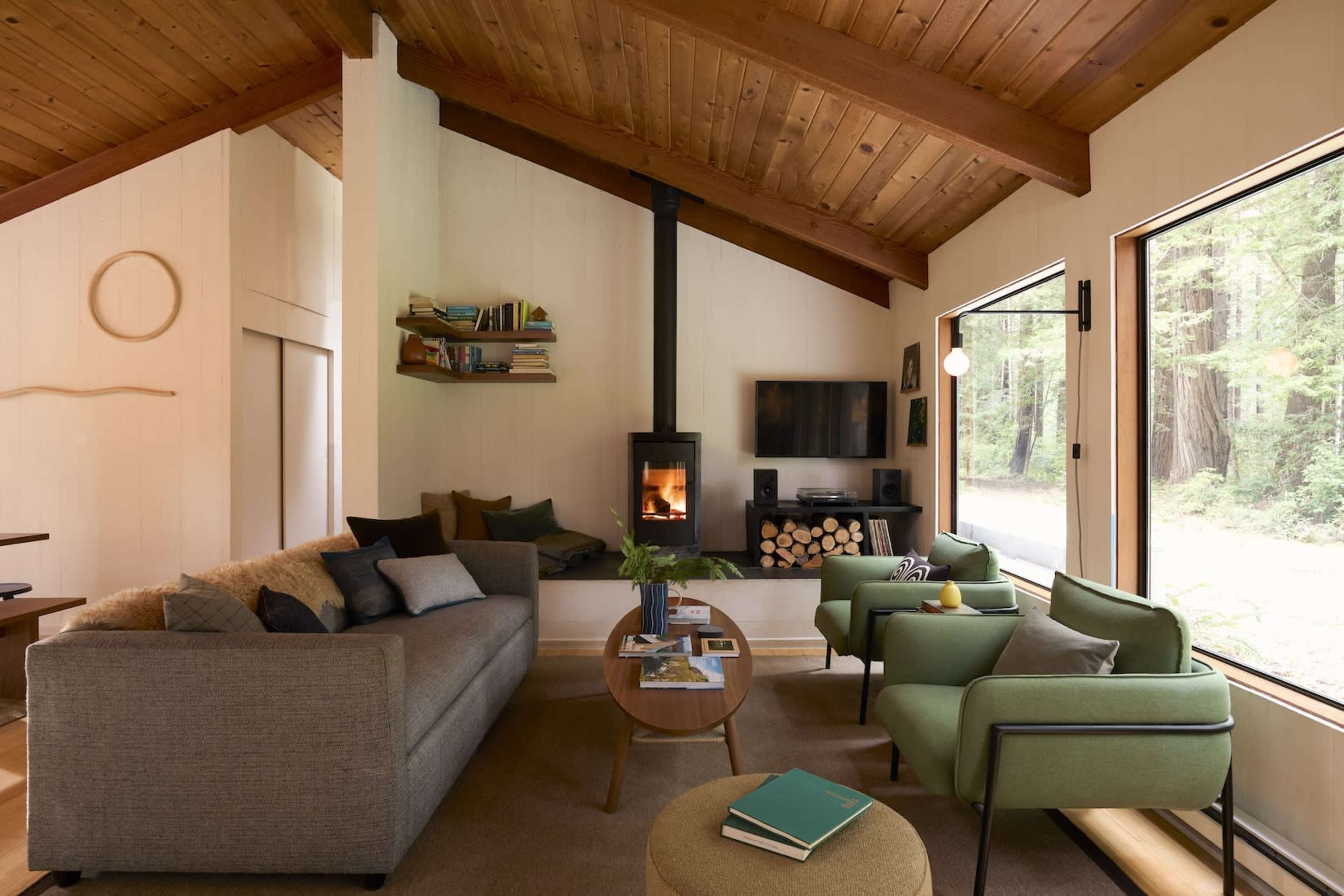 A modern living room with a gray sofa, two green chairs, a wooden table, a fireplace, and large windows overlooking a forest.