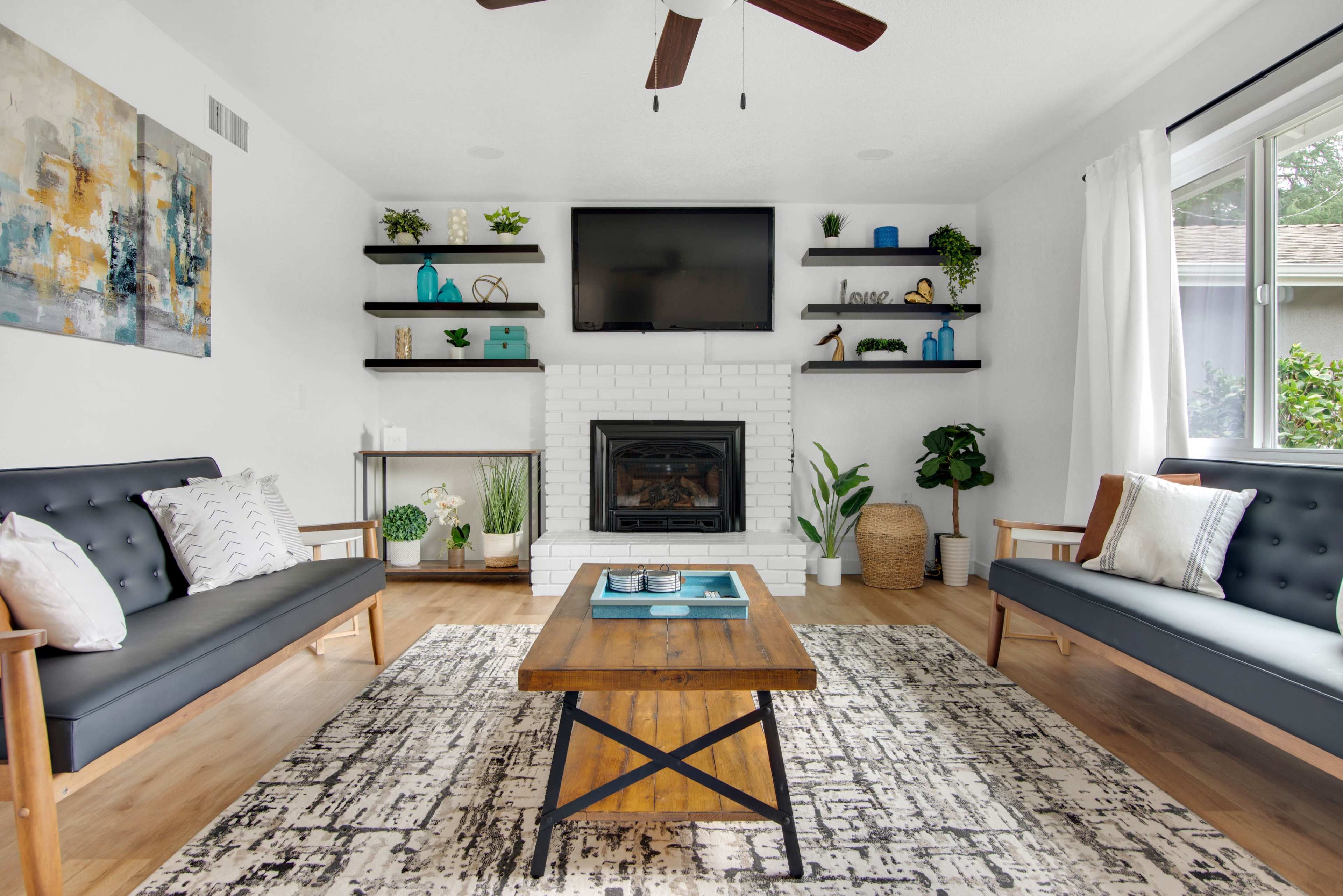 A modern living room features a white brick fireplace, a large flat-screen TV mounted above it, and two dark sofas flanking a wooden coffee table on a patterned rug.