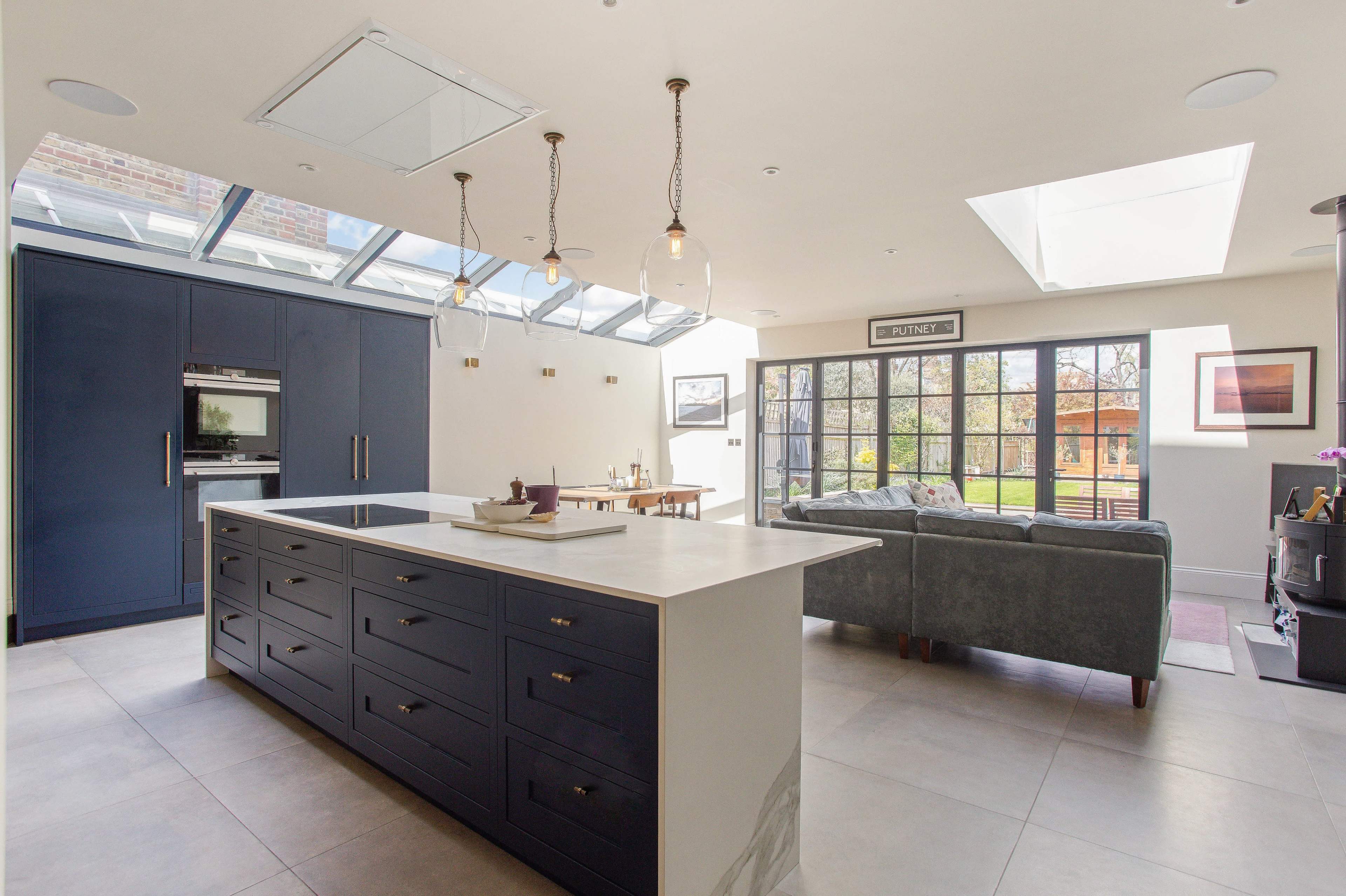 A modern kitchen features a central island with a white countertop, dark blue cabinetry, and large windows allowing natural light to fill the space.