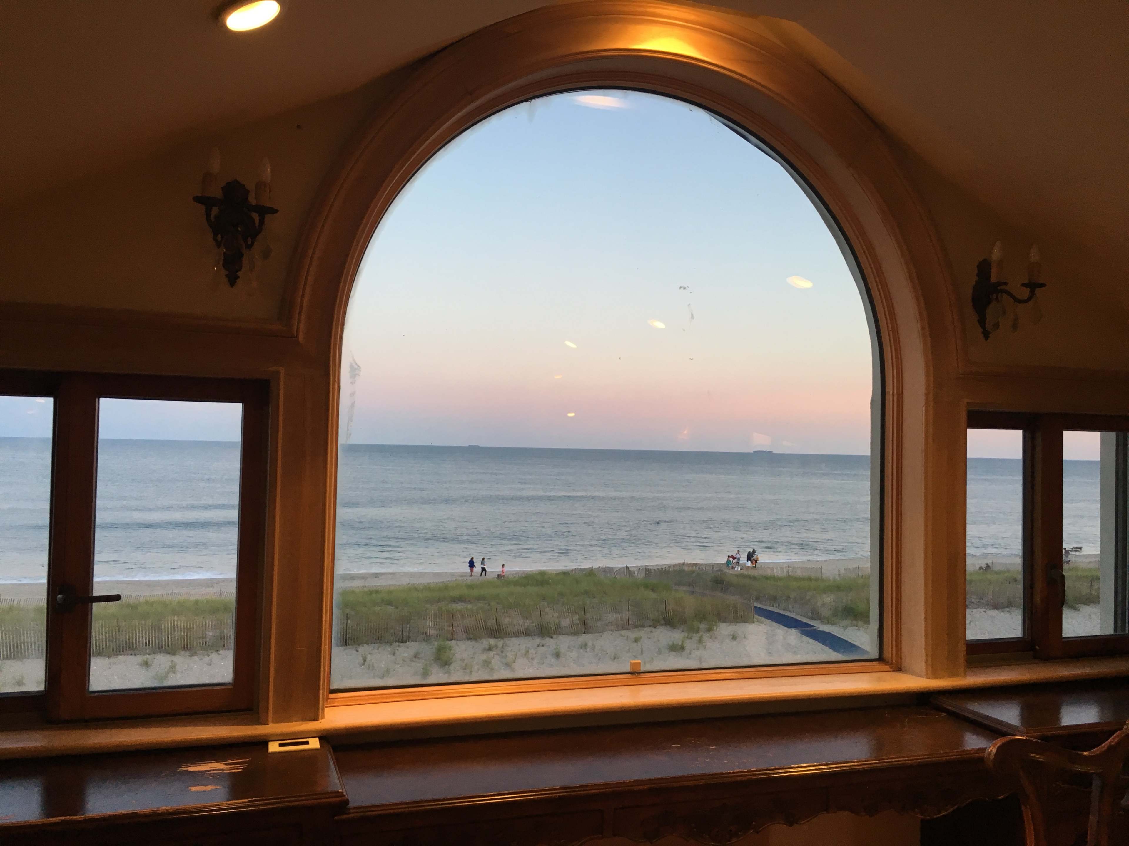 A large arched window overlooking a calm ocean with a sandy beach and small figures in the distance.