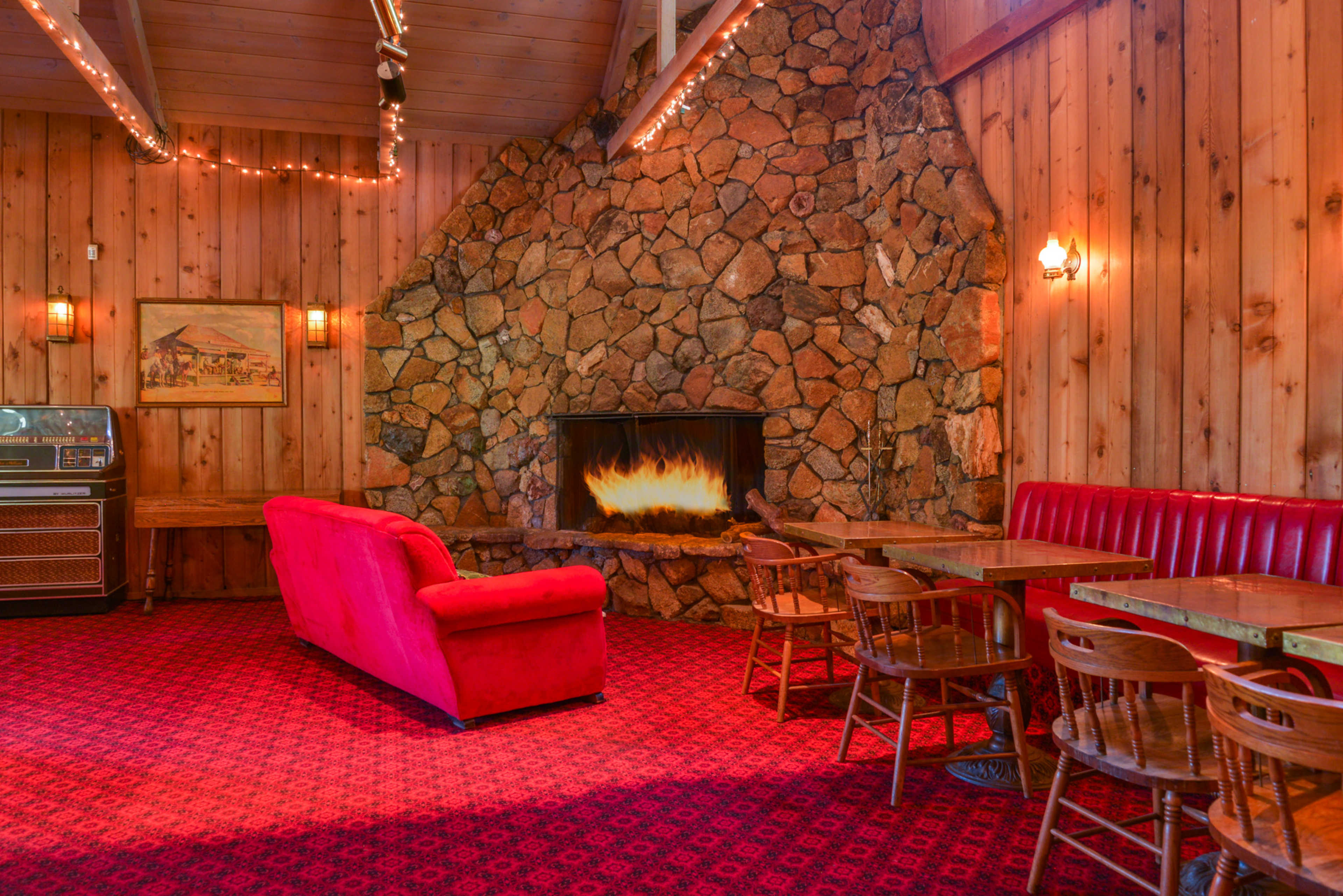 The scene features a cozy room with a stone fireplace, wooden walls, a red couch, and several wooden tables surrounded by chairs.