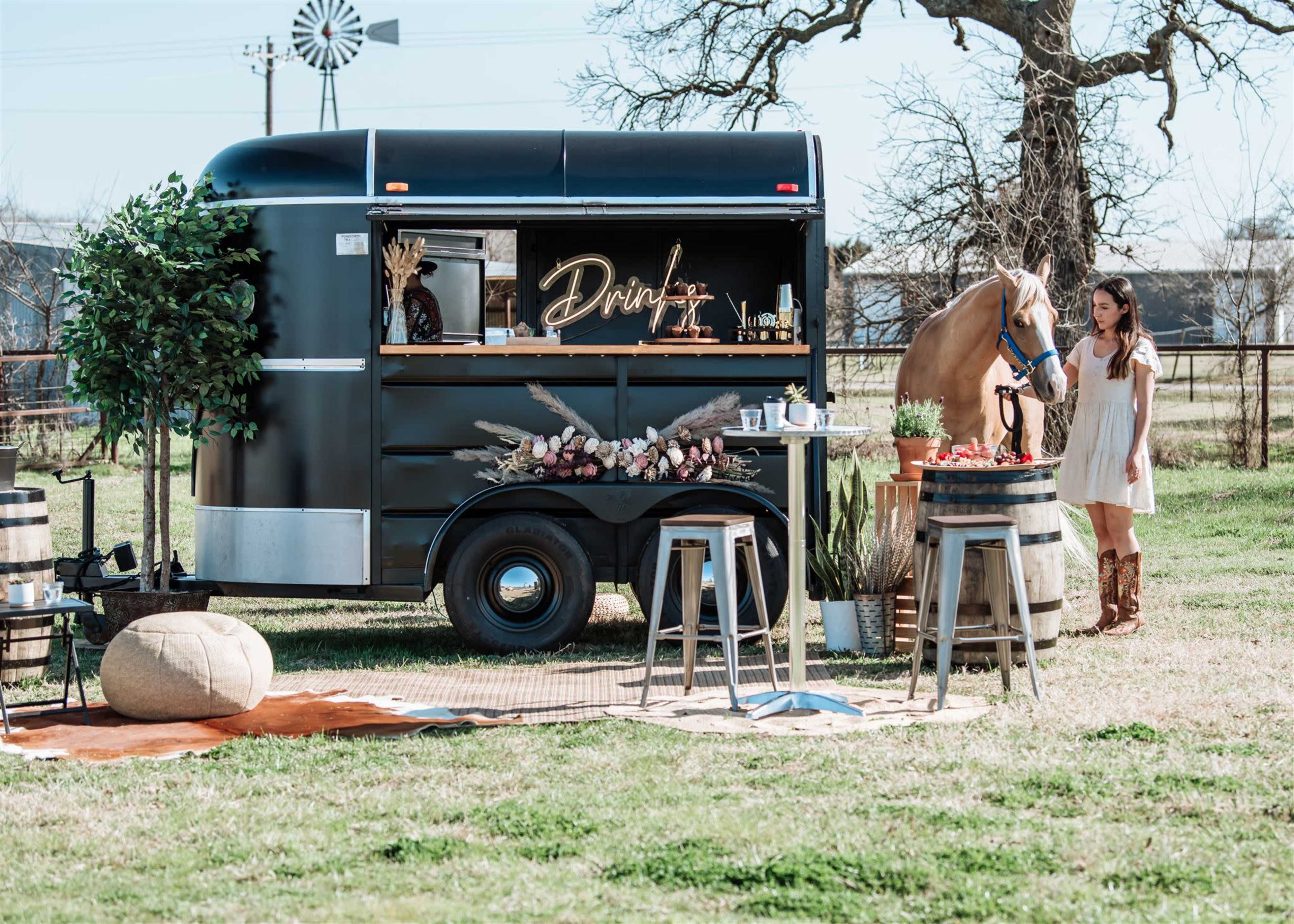 A woman stands next to a horse by a black beverage trailer set up in a grassy outdoor area with rustic decor and seating.